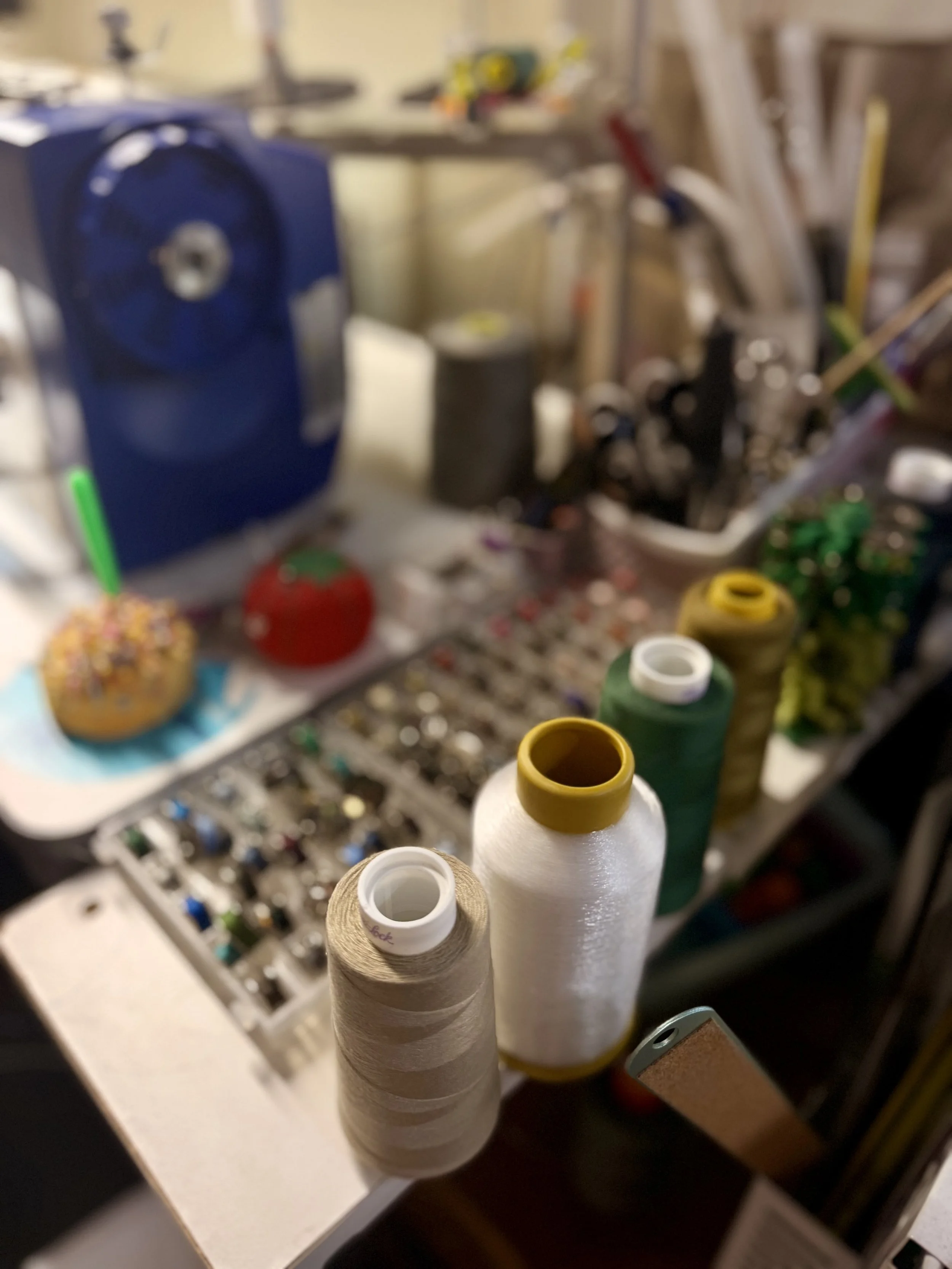 Several spools of thread in various colors are standing upright on a surface, with a blurry background of sewing supplies and tools.