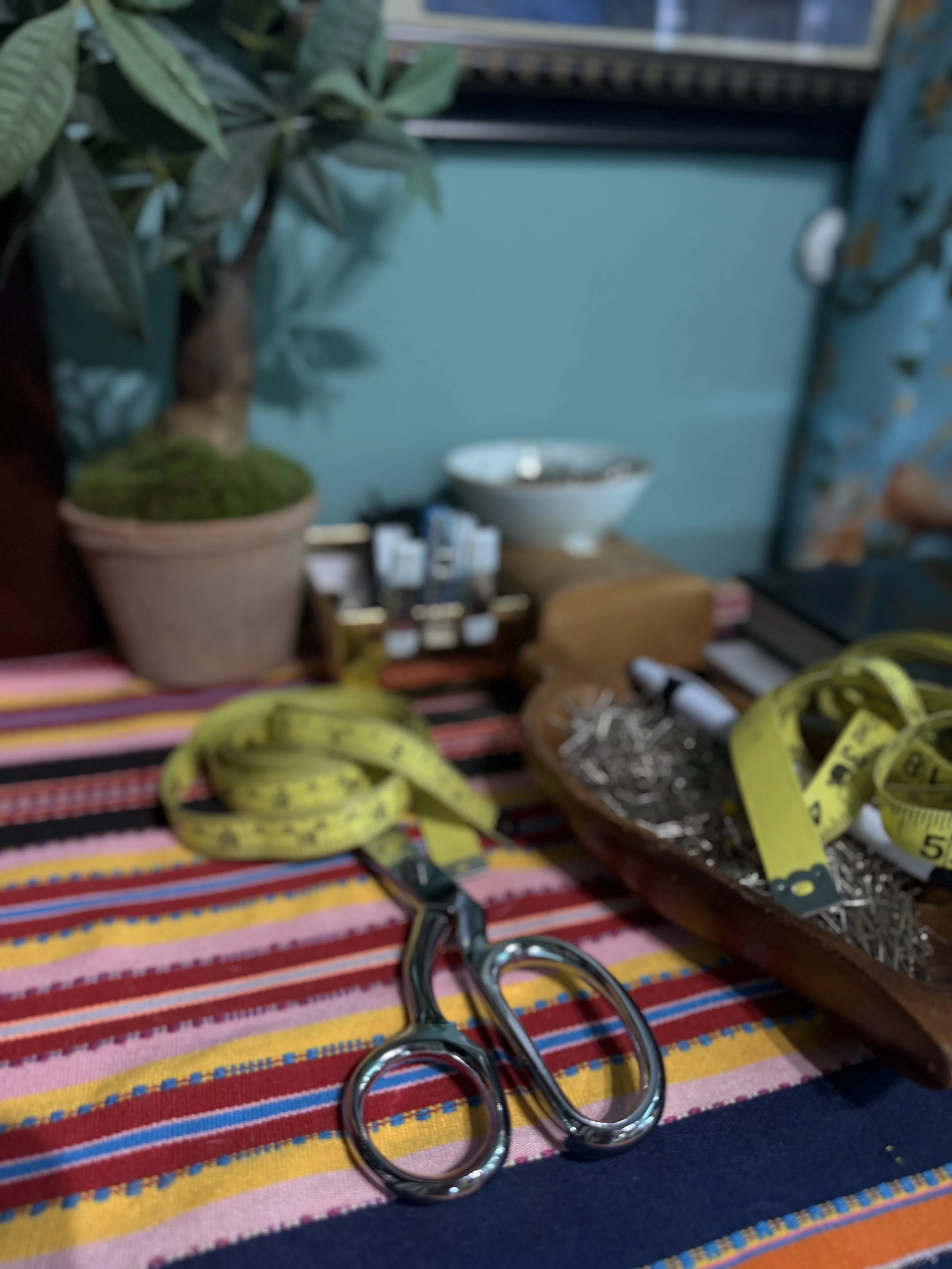 A colorful striped fabric surface with a pair of shiny scissors, a yellow measuring tape, a bowl filled with metal pins, and various small objects. In the background, there is a small potted plant with green leaves, a bowl, and some bottles, all placed on a wooden surface.