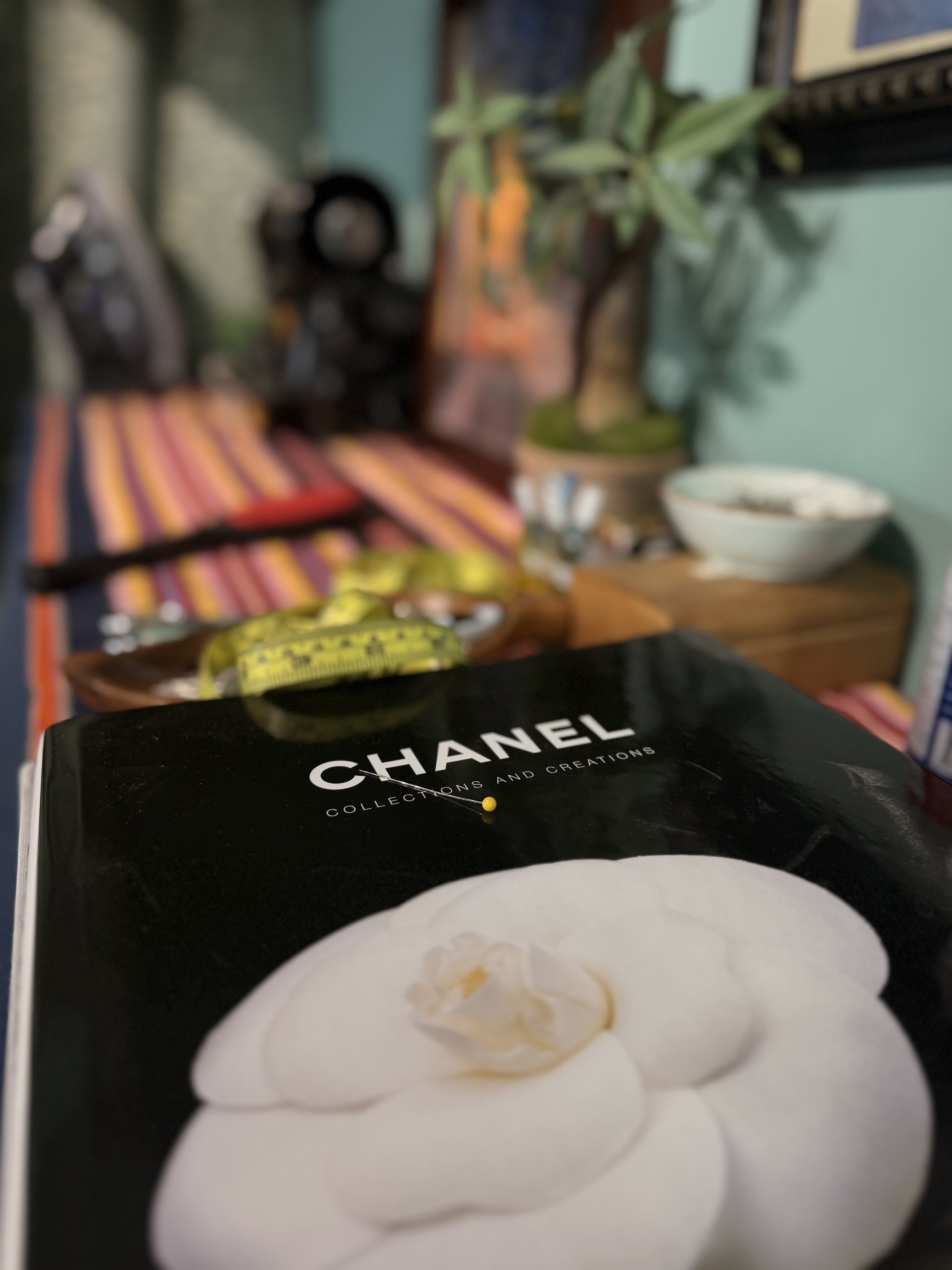 Close-up of a book of Chanel Collections and Creations in the foreground, with a blurred background showing a potted plant, bowl, and various objects on a shelf or table.