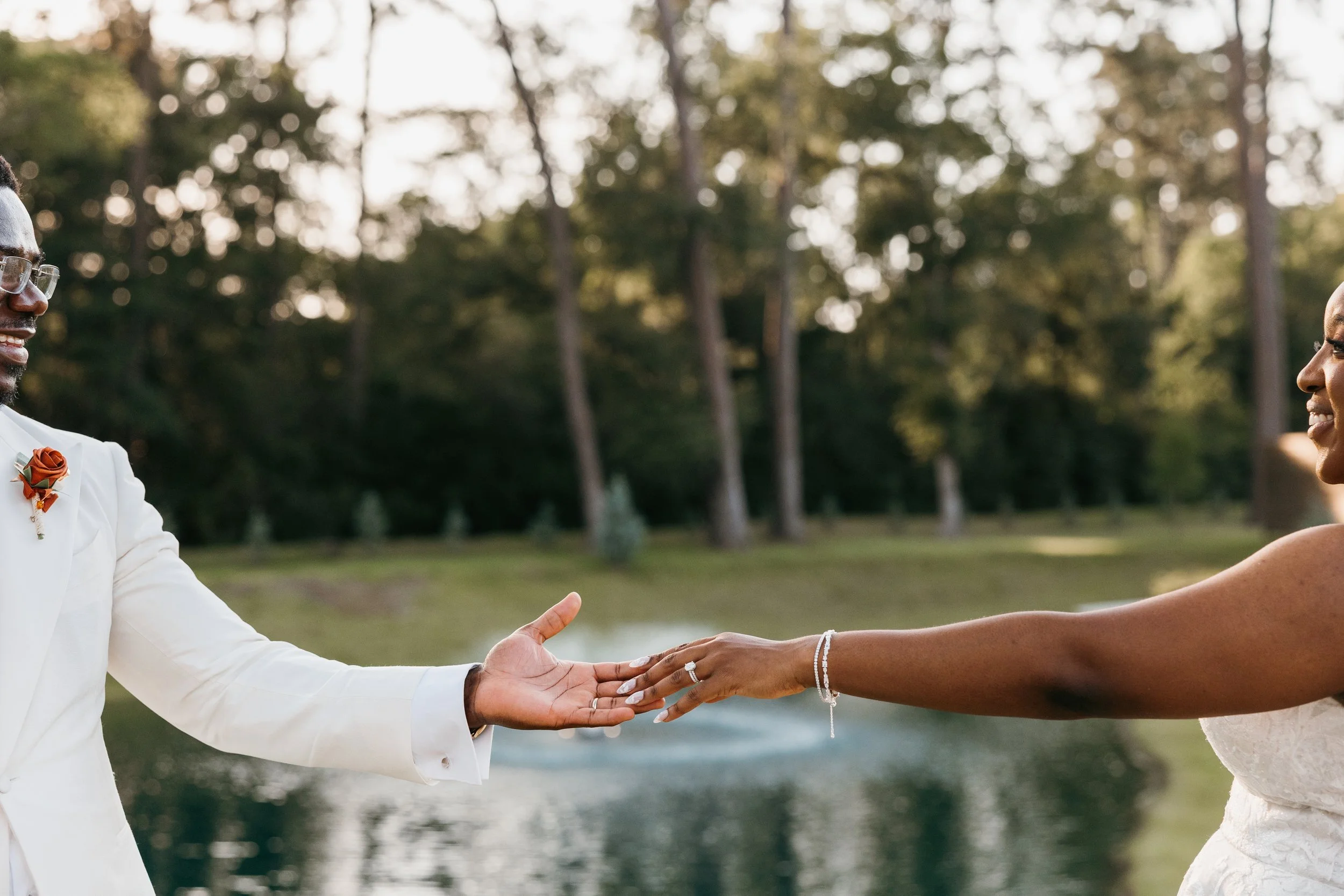 Couple holding hands outdoors, with the man wearing a white suit and boutonniere, and the woman in a white dress, during sunset in a park with trees in the background.