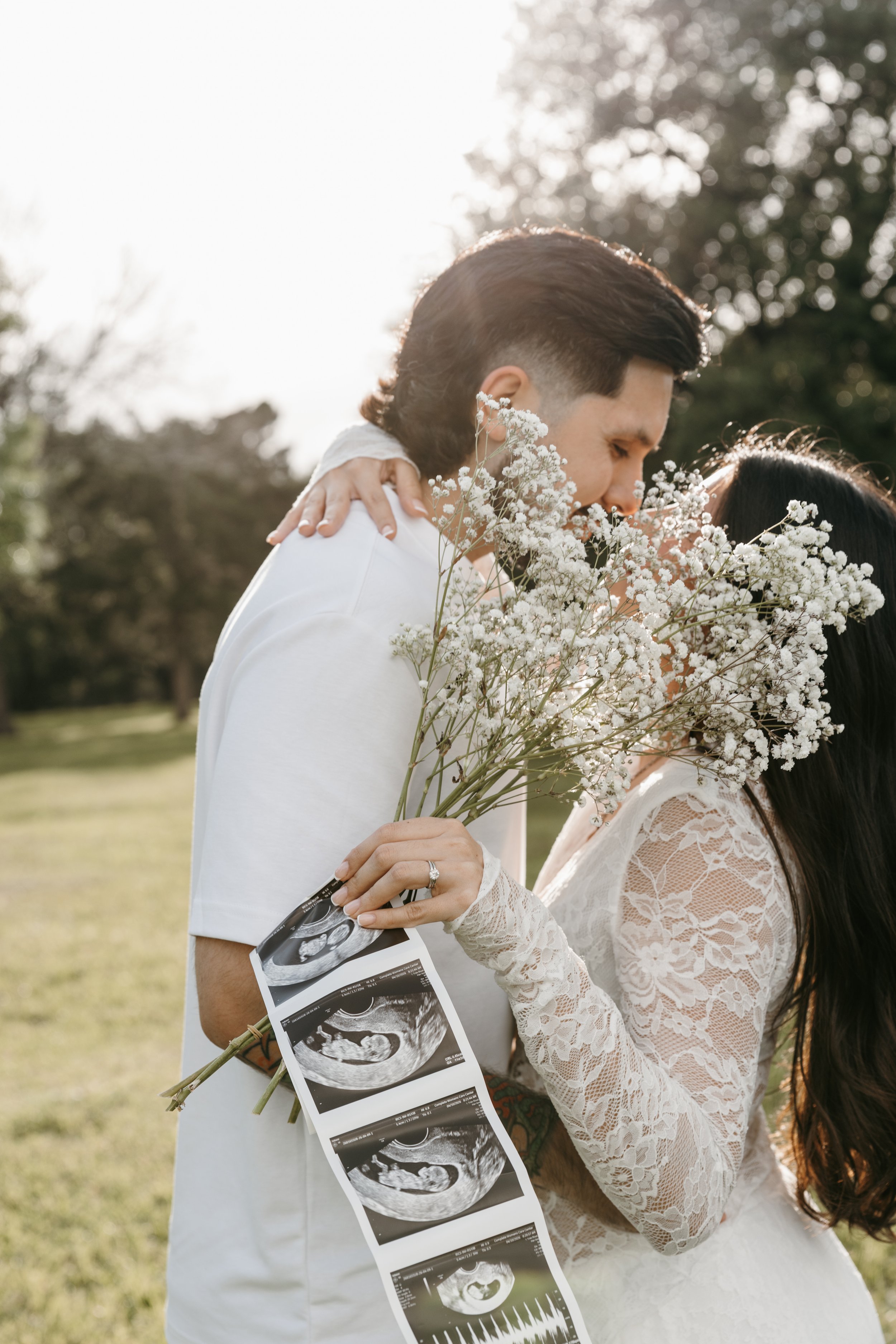 A couple at a park during a sunny day, with the man holding ultrasound pictures and a bouquet of white flowers, the woman wearing a lace dress, and they are close together with faces touching.