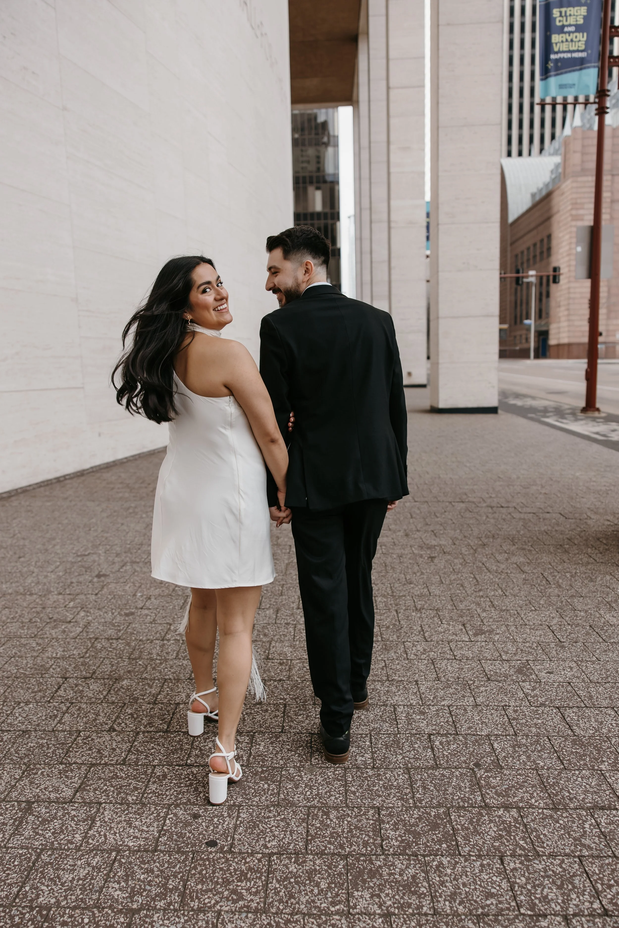 A smiling woman in a white dress and high heel shoes holding hands with a man in a black suit walking in an urban area with modern buildings.