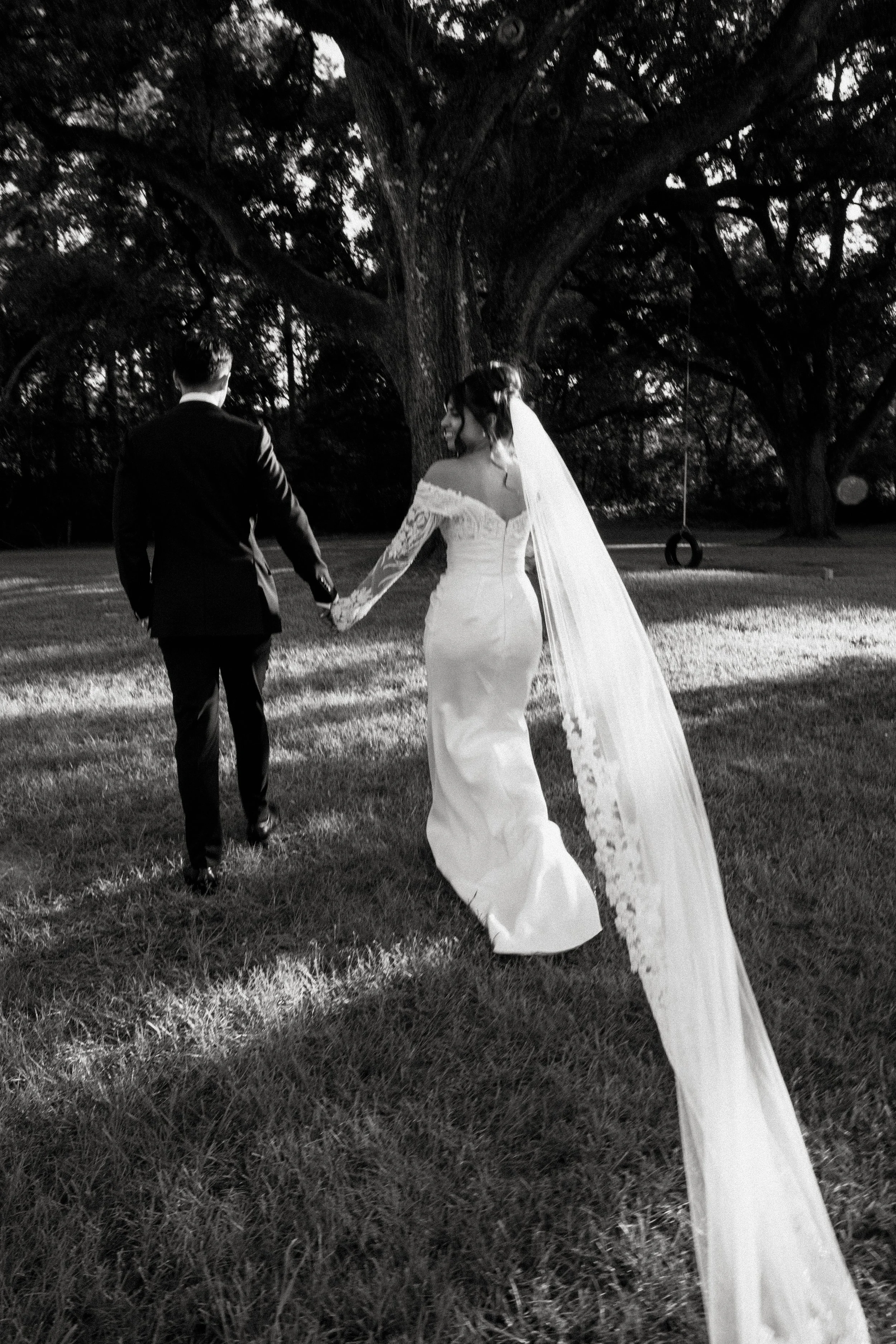 A bride and groom holding hands walking outdoors near a large tree, with a tire swing hanging from the branches, during dusk.