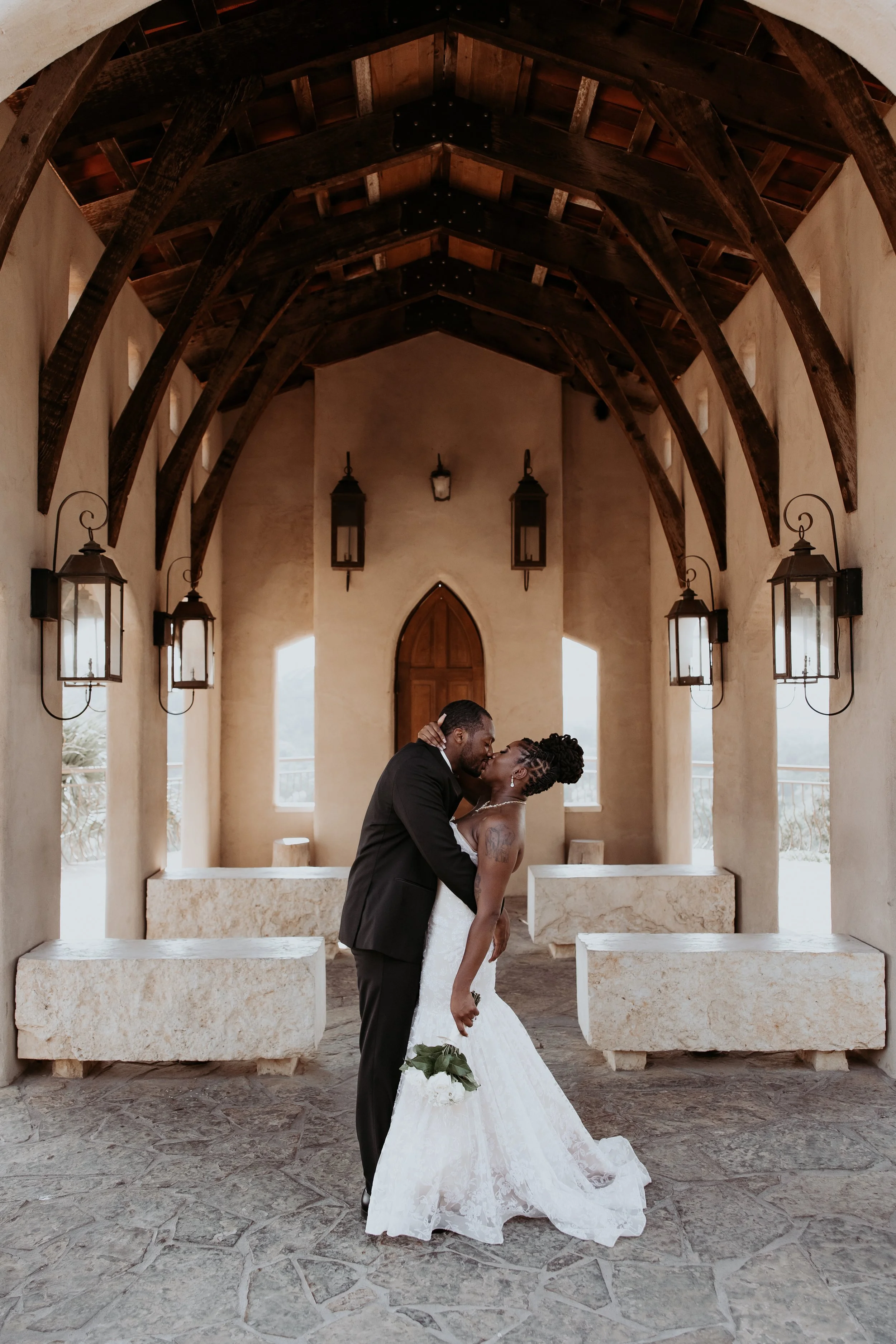 An engaged couple sharing a kiss in a beautifully lit, enclosed outdoor space with wooden beams and lanterns on the walls.