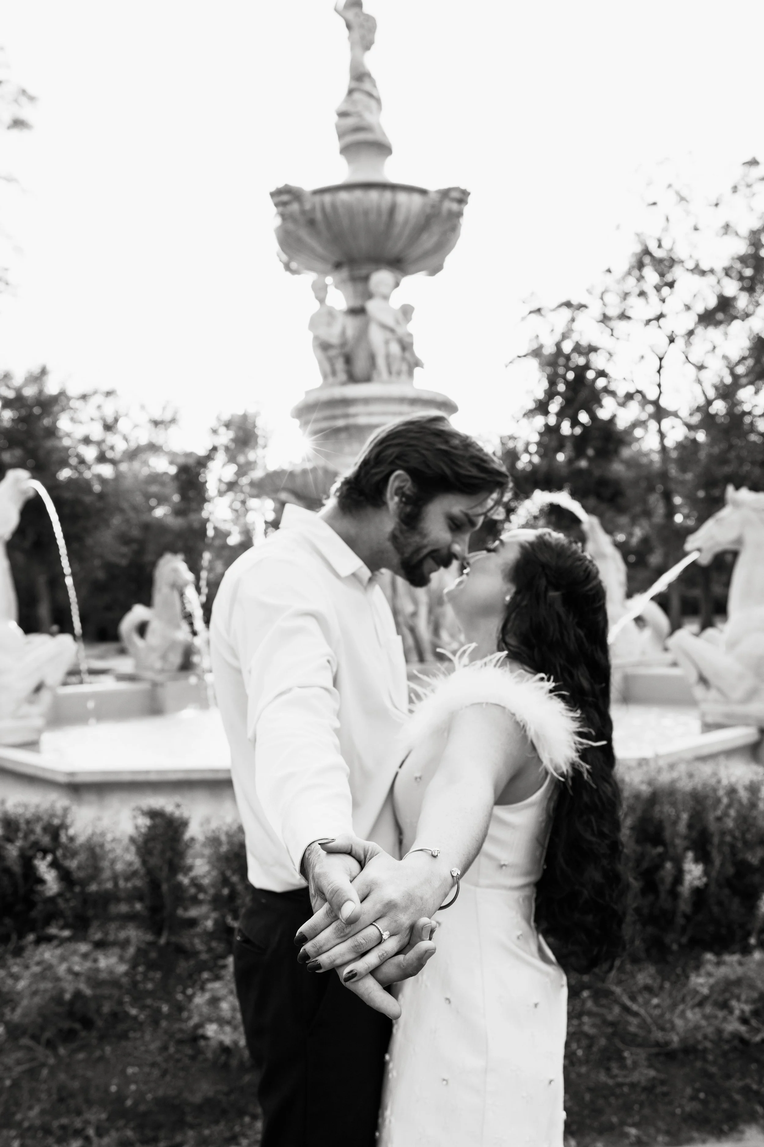 A black and white photo of a couple dancing outdoors near a fountain with sculptures, trees, and a bright sky in the background.