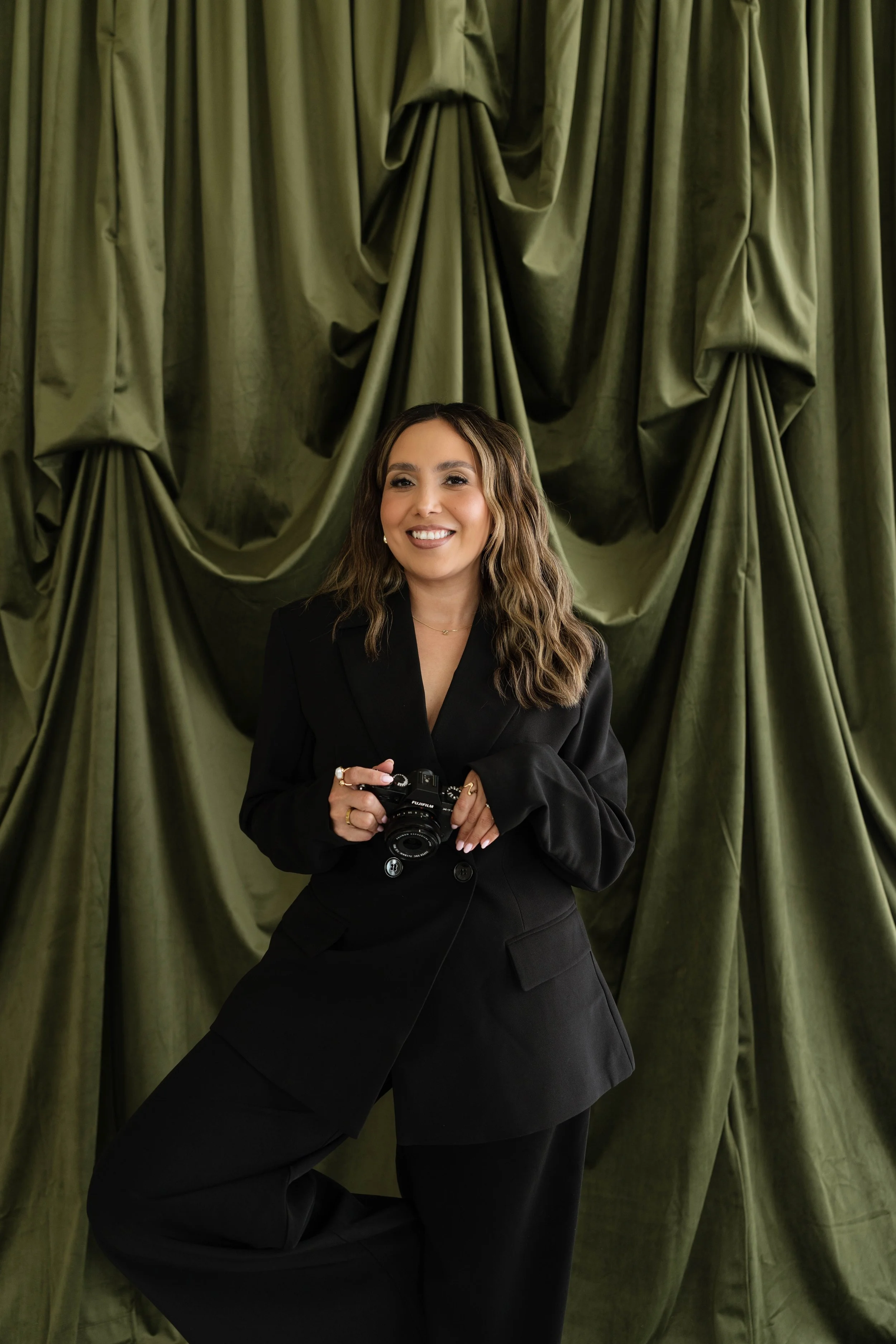 A woman smiling while holding a camera, standing in front of green draped curtains.