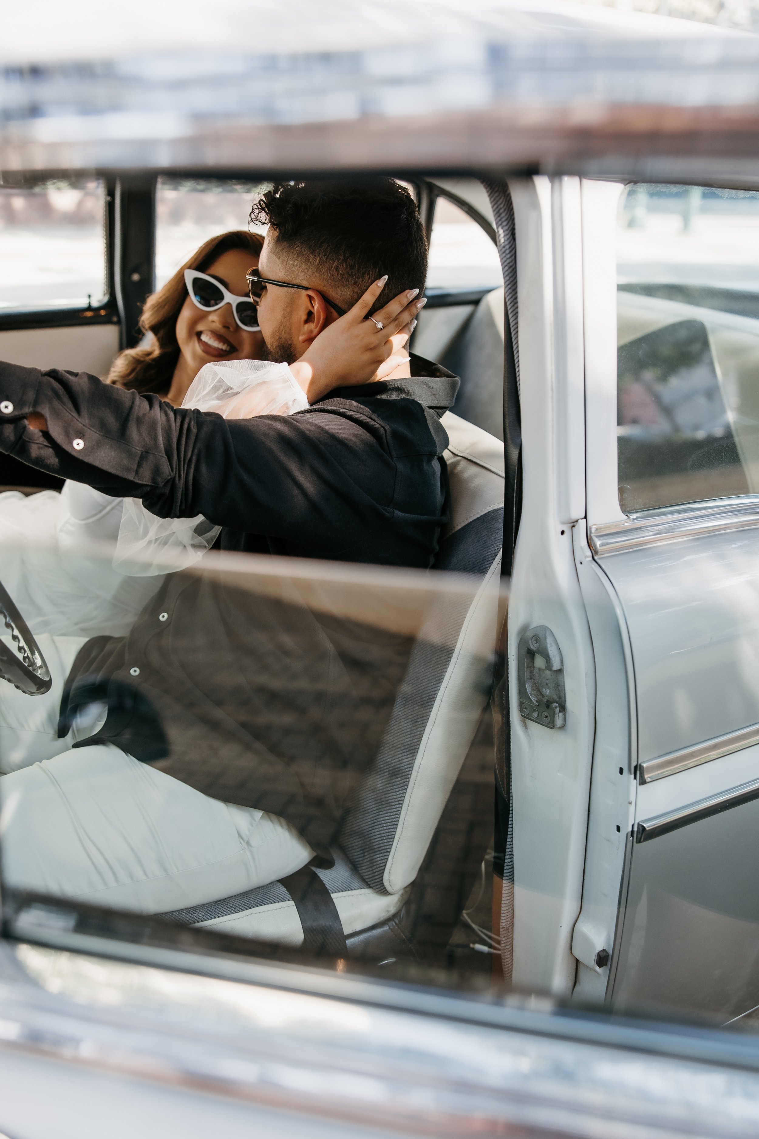 A couple sitting in a vintage car, sharing a joyful moment, with the woman touching the man's face and both laughing. The woman is wearing white sunglasses and a white outfit, while the man is wearing black sunglasses and a black jacket.