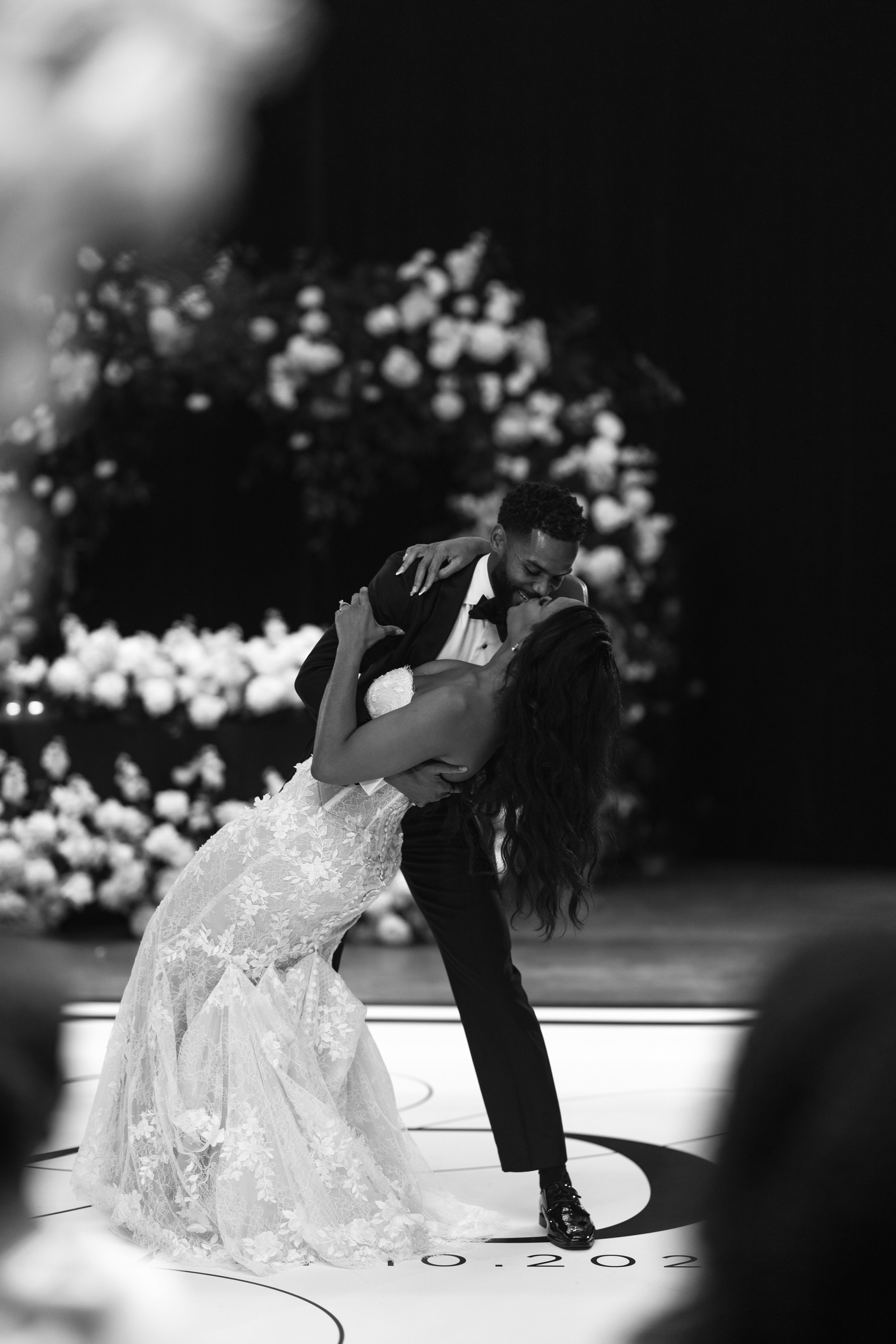 A black and white photo of a couple dancing, with the man in a tuxedo and the woman in a wedding dress, on a stage decorated with flowers.