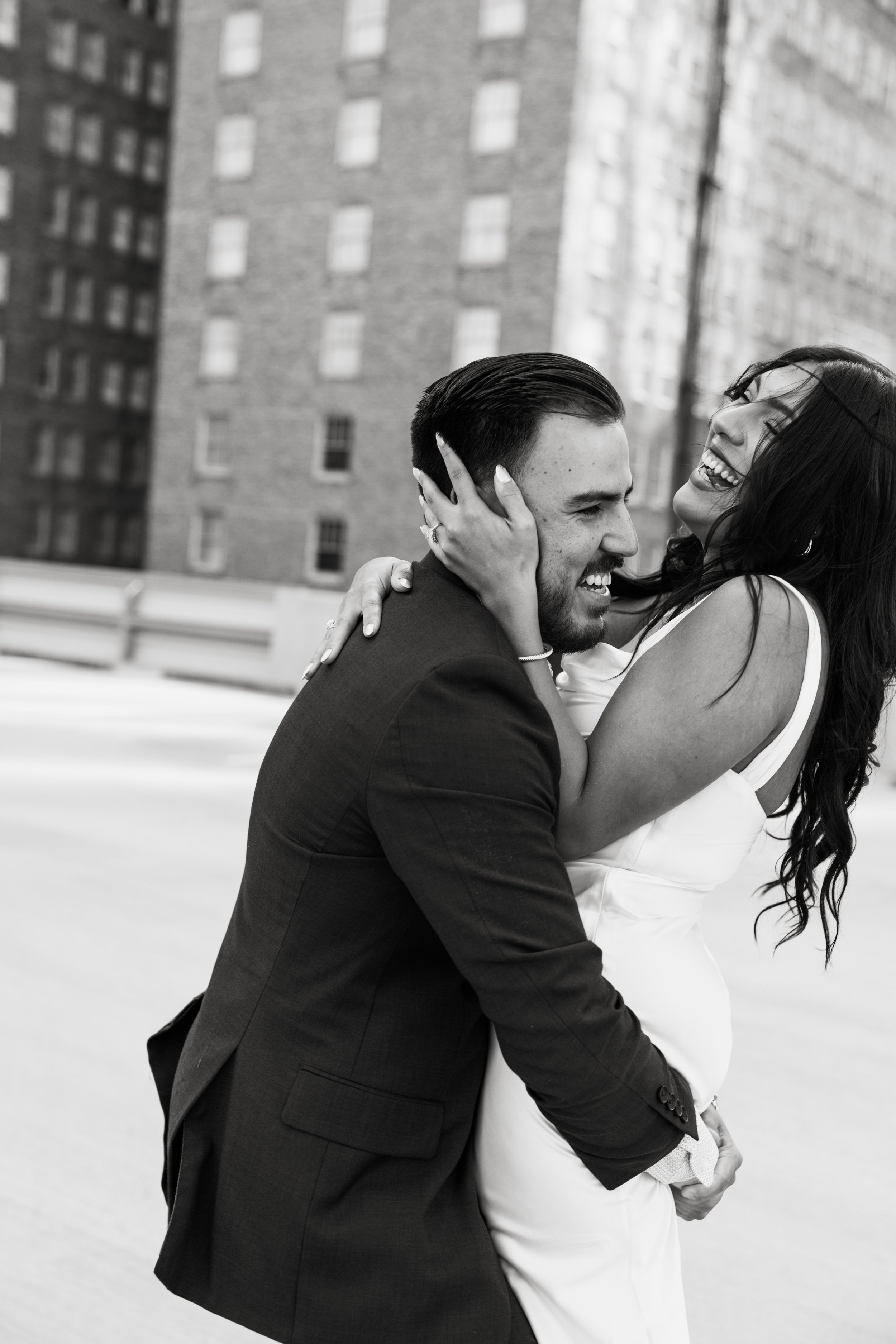 Black and white photo of a man and woman embracing and laughing on a rooftop with a cityscape of brick buildings in the background.