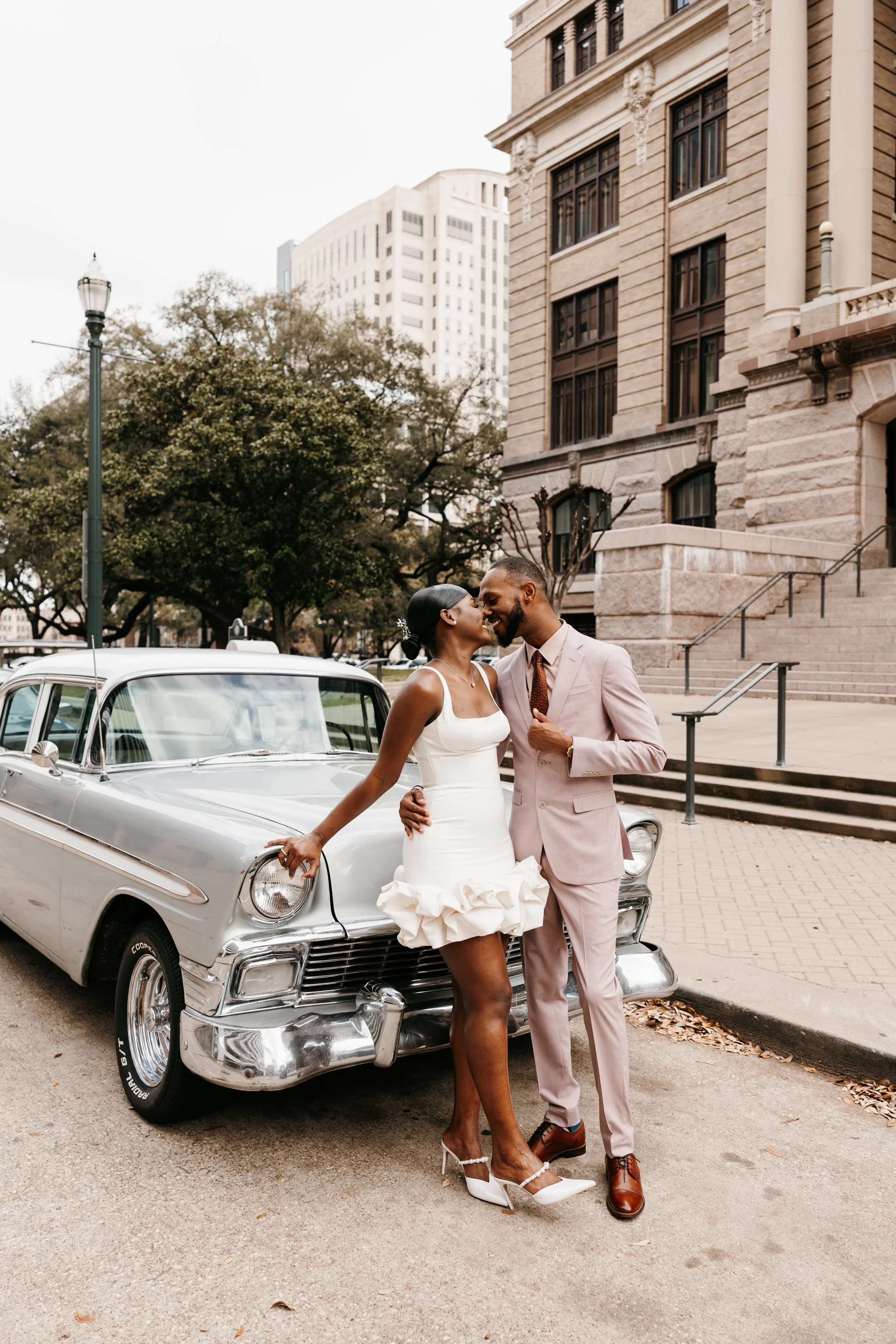 A couple dressed in wedding attire standing next to a vintage silver car on a city street. The woman is wearing a white wedding dress and high heels, and the man is wearing a light pink suit with a brown tie. They are smiling and touching foreheads. 