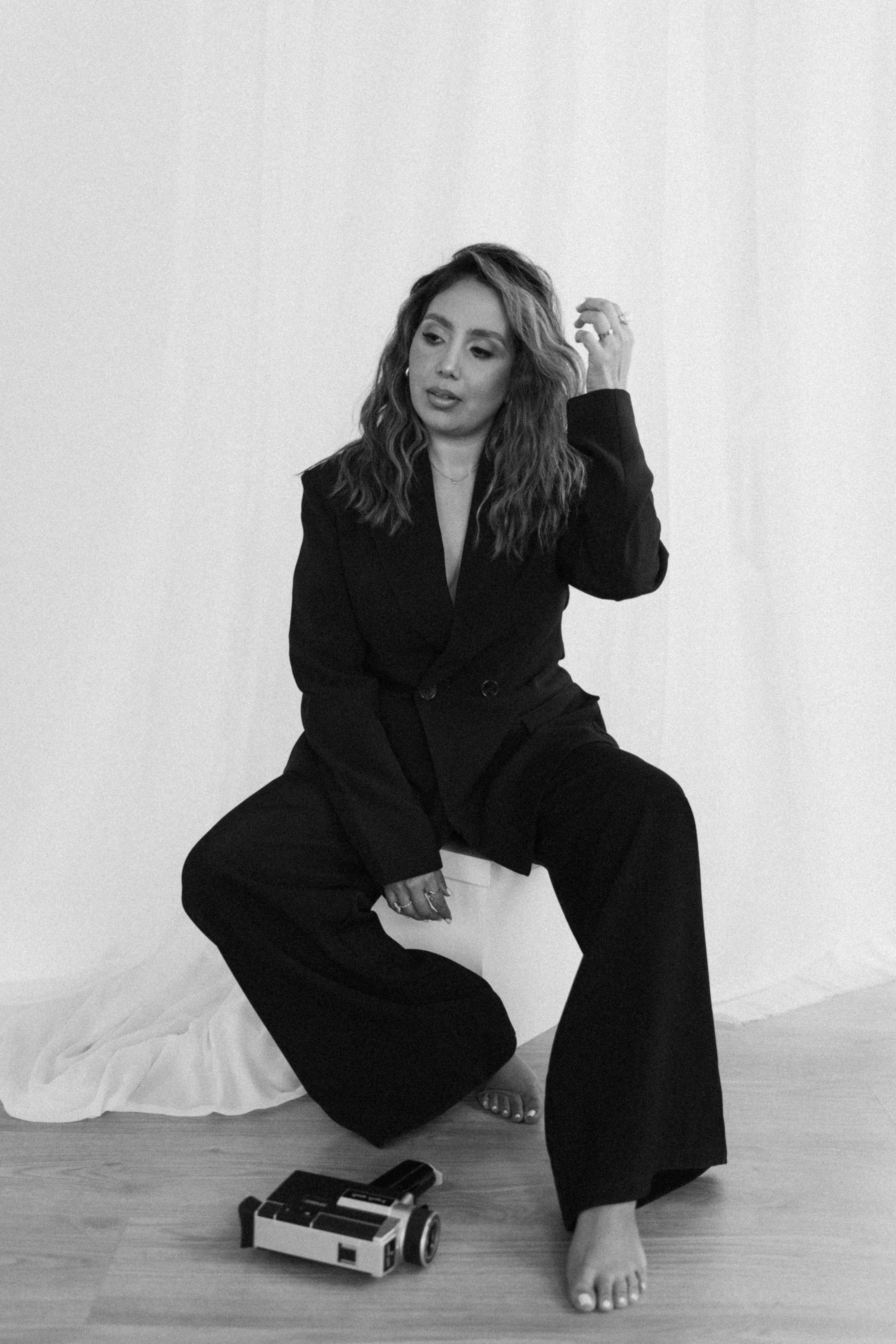 Black and white photo of a woman sitting on the floor in front of a plain wall. She has wavy hair and is wearing a dark blazer and wide-leg pants. Her left hand is touching her hair, and her right hand is resting on her knee. There is a toy robot on the floor in front of her, and a piece of cloth or sheet on the floor near her feet.