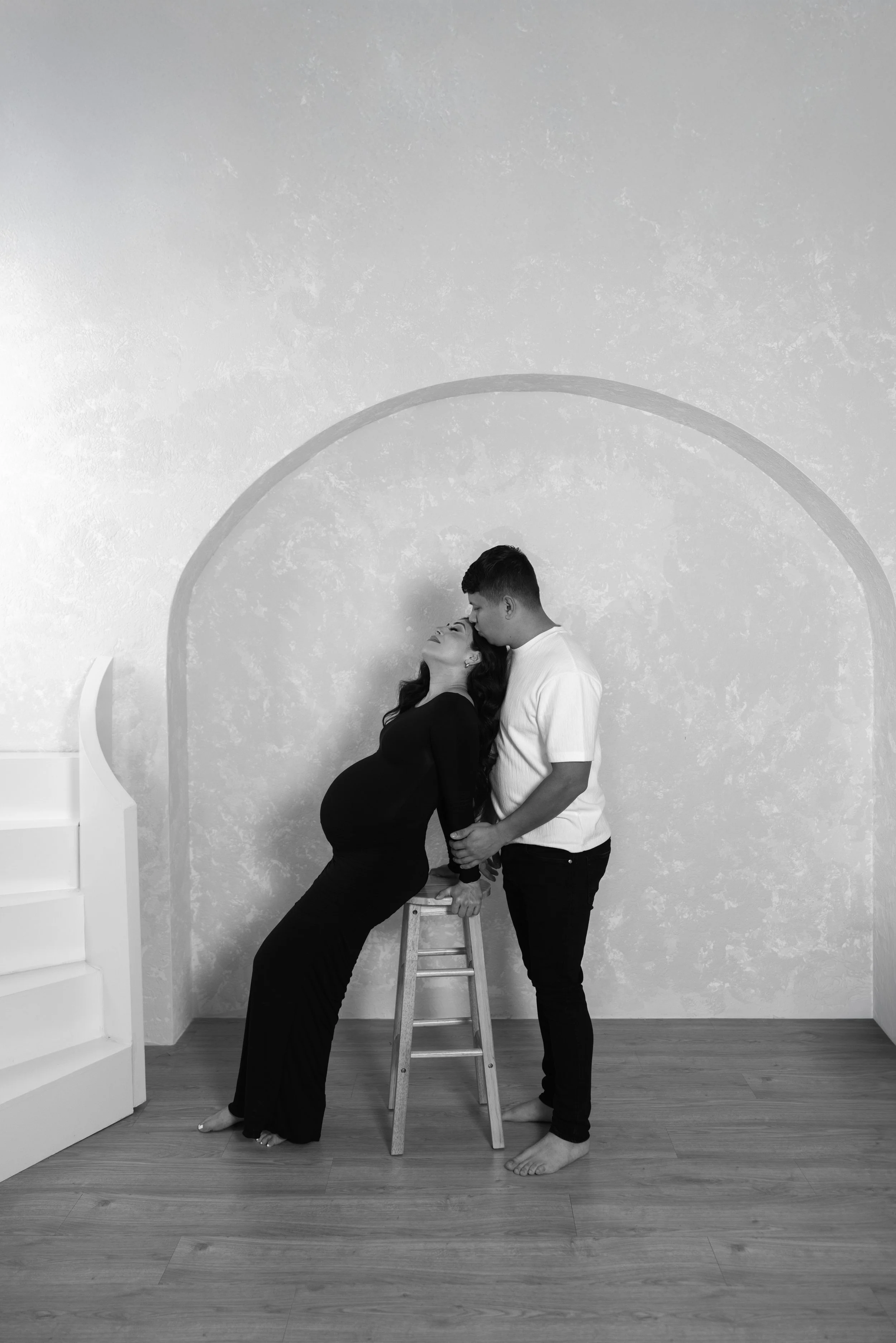 A pregnant woman and a man standing behind a stool, with the woman leaning back and the man gently holding her waist, in a room with a textured wall. The woman is wearing a black dress and the man a white shirt and dark pants.