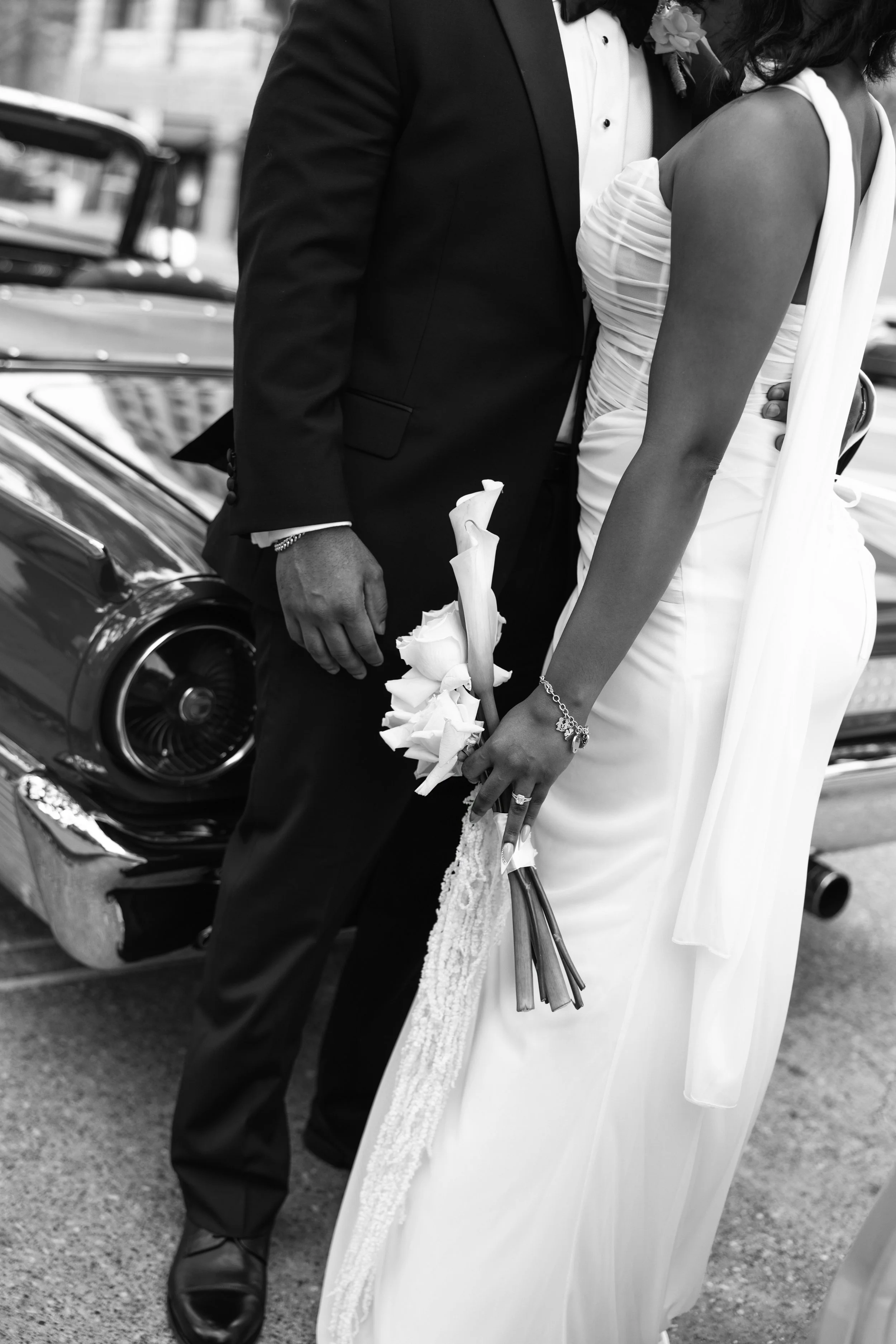 Black and white close-up of a bride and groom at their wedding, standing near a vintage car. The groom is wearing a tuxedo and holding a boutonniere, while the bride is in a white gown holding a bouquet of flowers. The focus is on their torsos and hands.