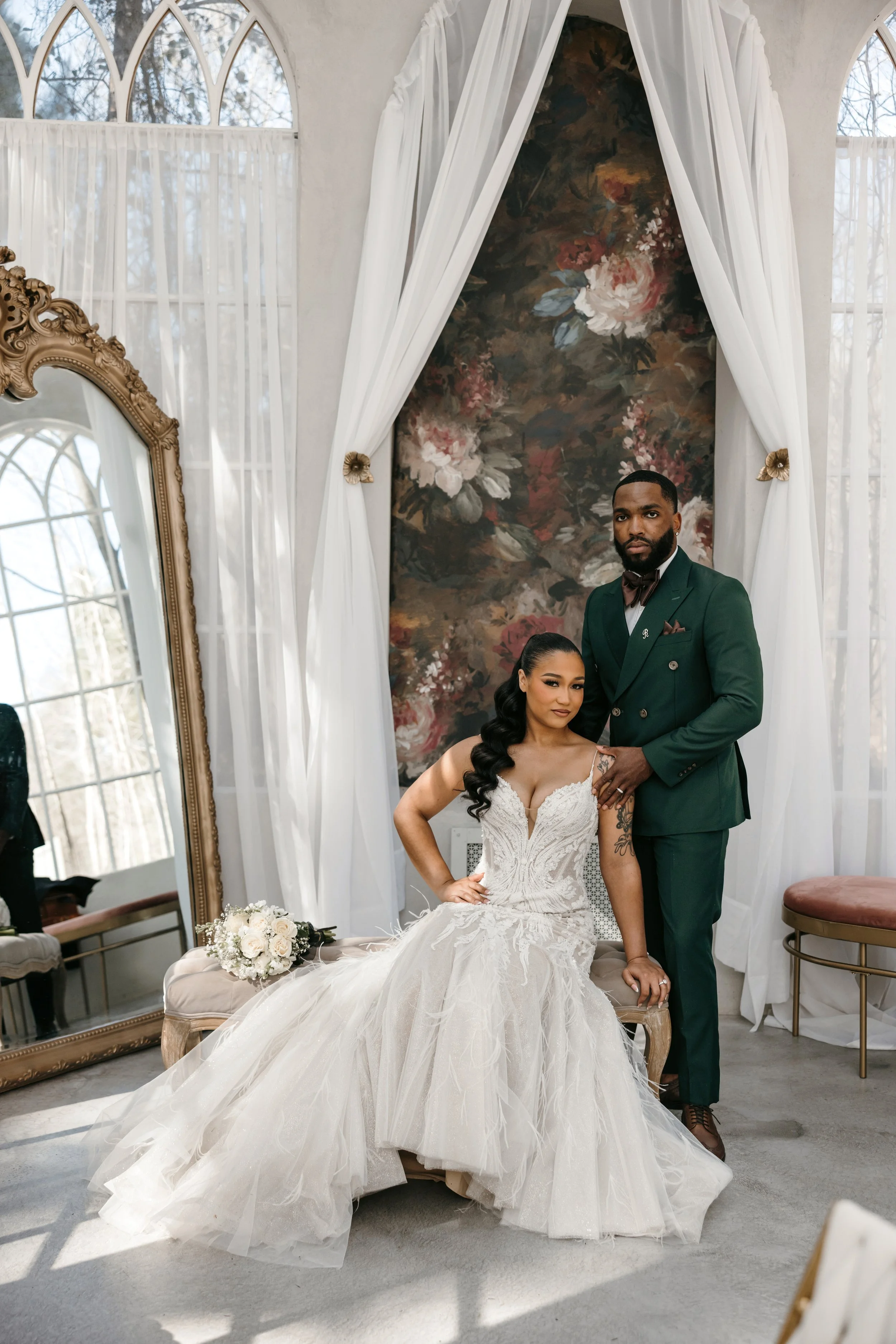 A bride in a white lace wedding gown sitting on a vintage-style bench, and a groom in a green suit with a bow tie standing behind her, in a bright room with large arched windows, white curtains, a floral backdrop, and a gold-framed mirror.