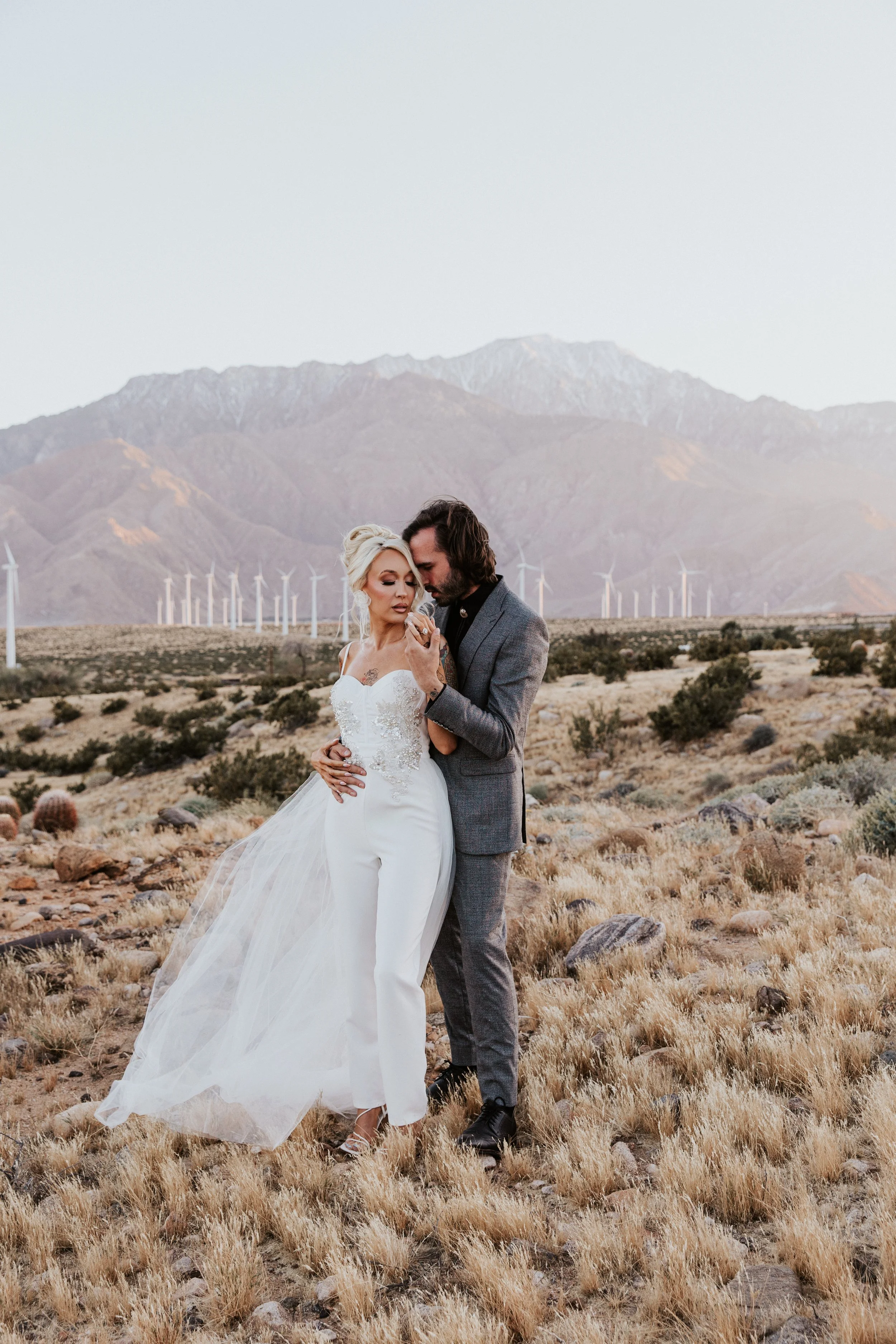 A bride and groom embracing in a desert landscape with mountains and wind turbines in the background.