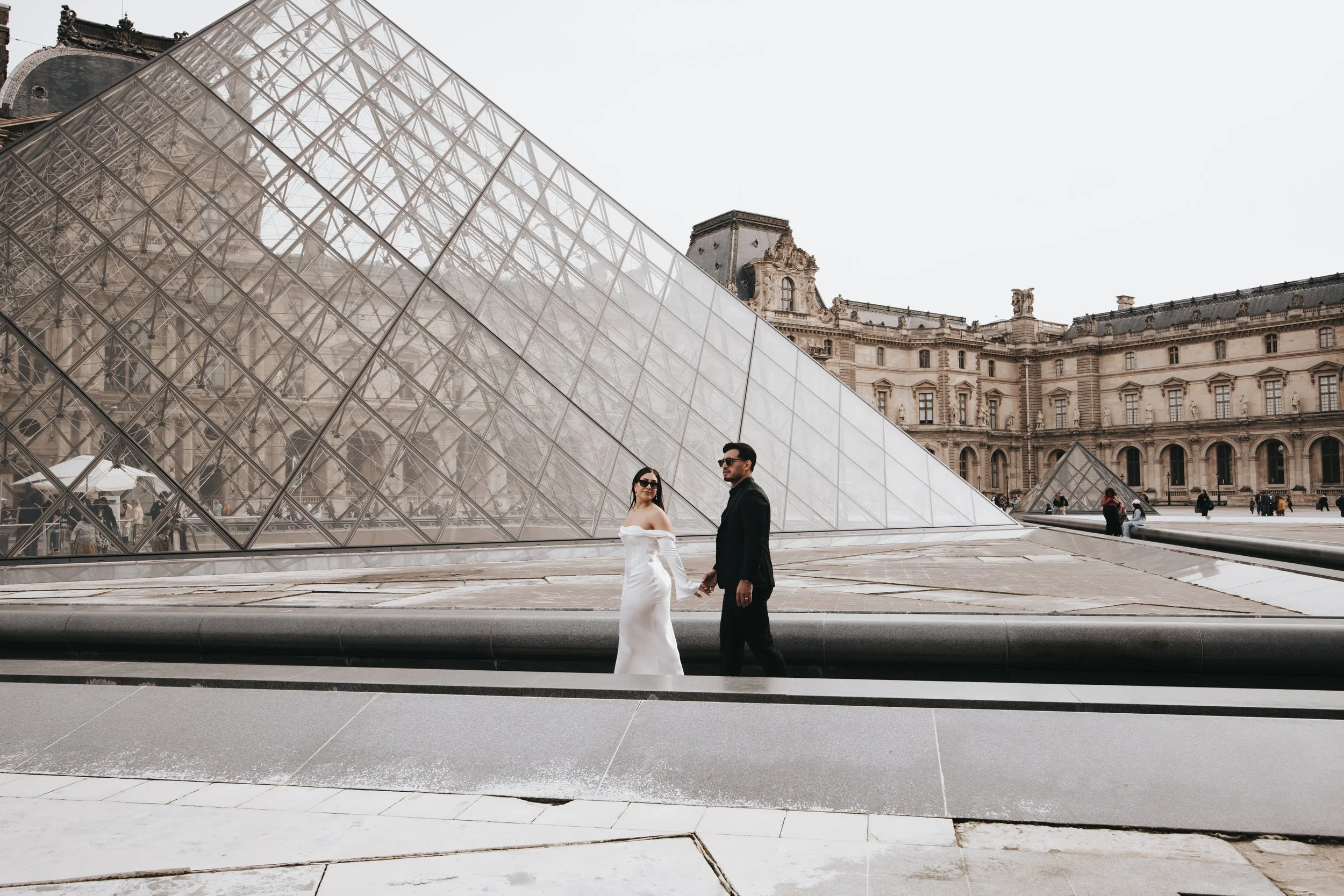 A couple dressed in wedding attire, holding hands, standing outside the Louvre Museum in Paris, France. The glass pyramid entrance and historic building are visible in the background.