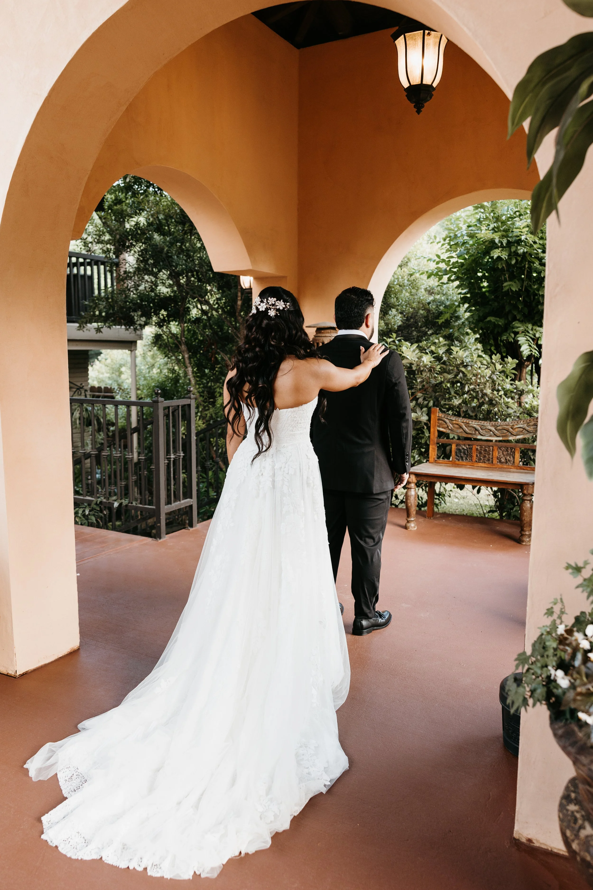 A bride in a white wedding dress with a long train and a groom in a black suit are entering a covered outdoor area with peach-colored walls and arches. The bride has dark, wavy hair with a floral hairpiece, and the groom has short dark hair. There is