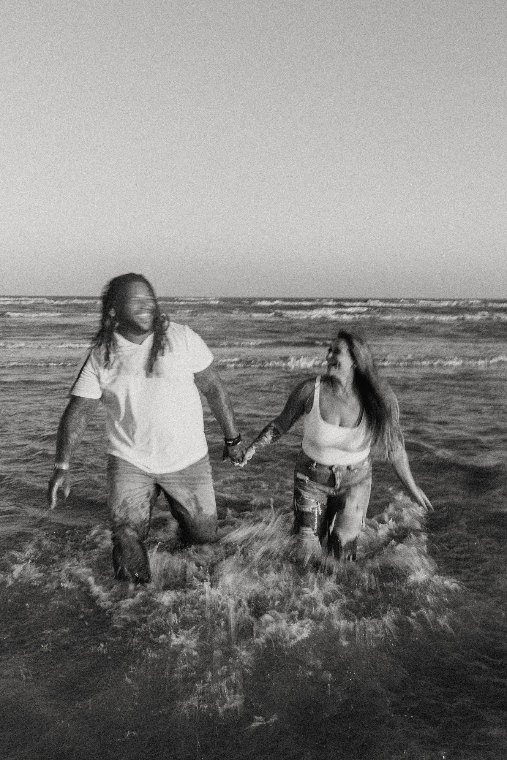 A black and white photo of a man and woman holding hands and running in the ocean at the beach. The man has long dreadlocks and tattoos, wearing a white t-shirt and shorts. The woman has long hair, wearing a tank top and ripped jeans. Waves are crash
