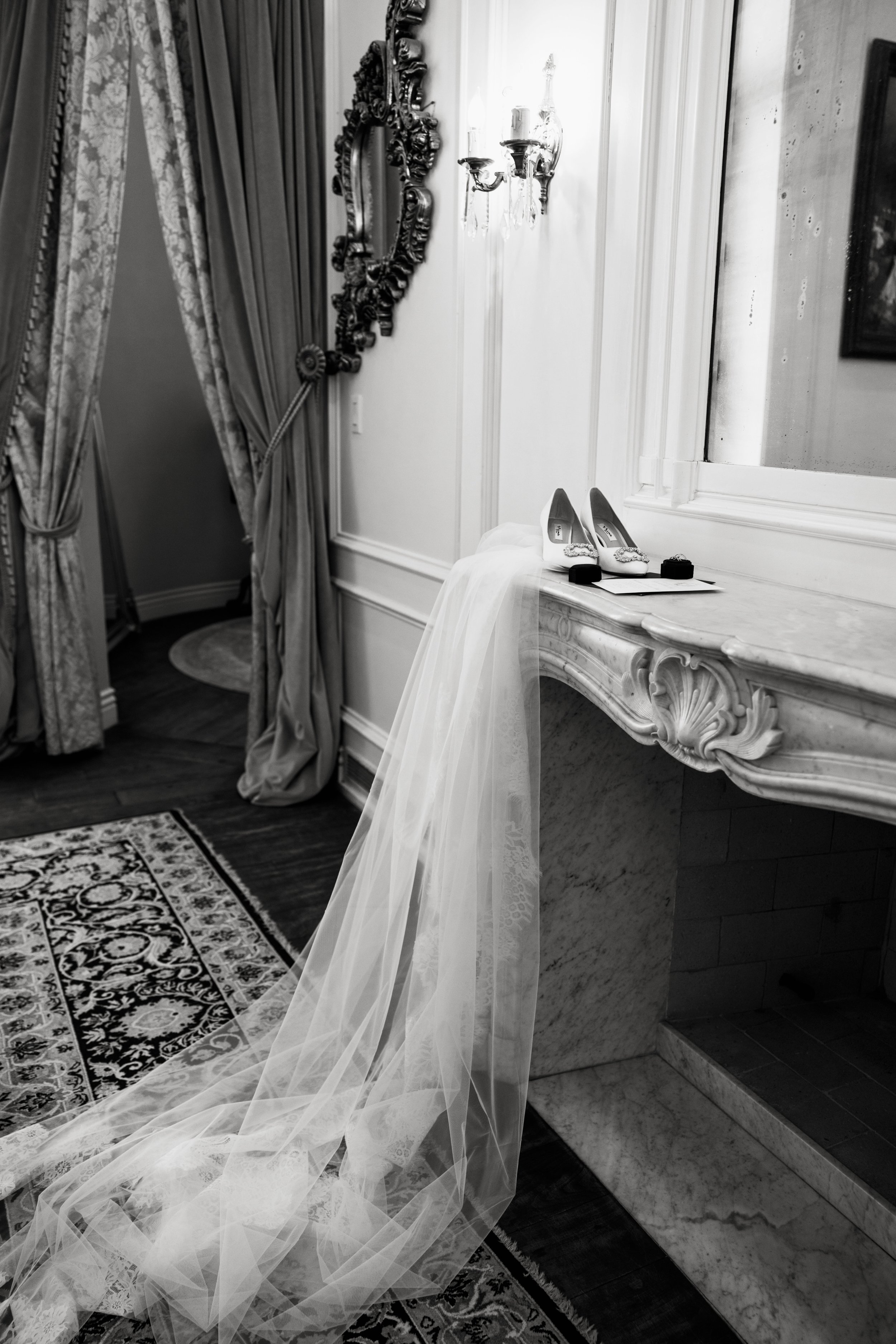 Elegant interior with ornate mirror, wall-mounted candle holder, pair of white high-heeled shoes on a marble vanity, sheer veil draping from the vanity, patterned rug on dark wood floor, decorated curtains in the background.