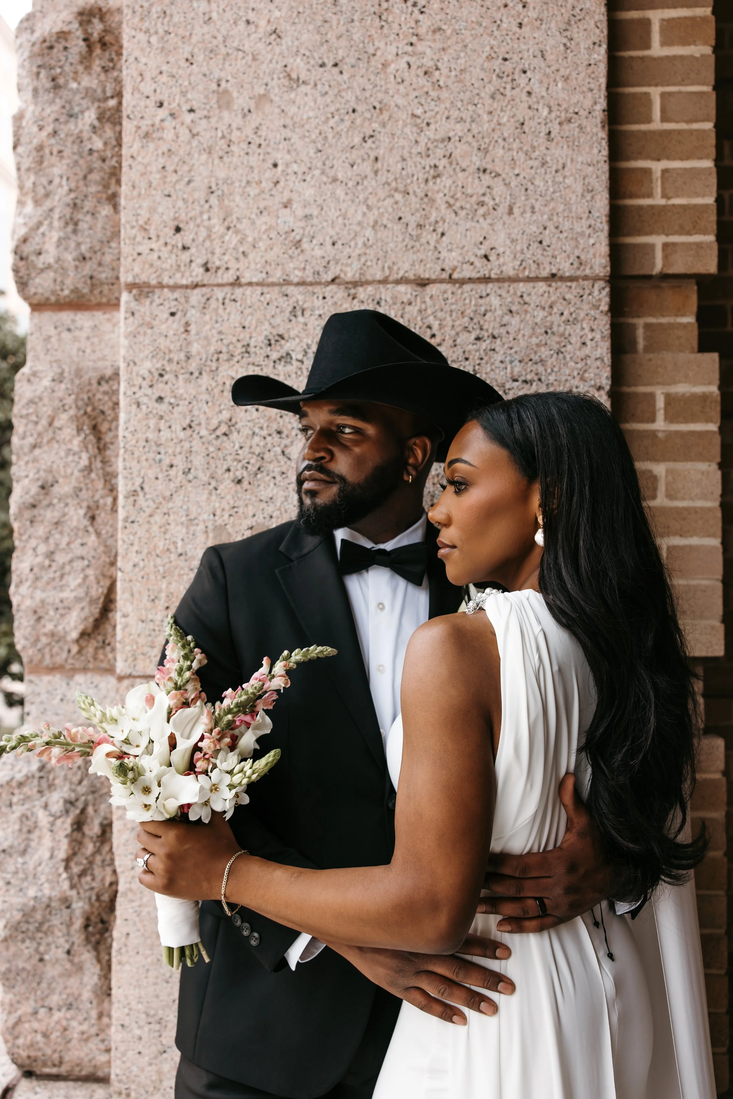 A couple dressed in formal wedding attire, standing close together outside near a brick and stone wall. The man, wearing a black suit and cowboy hat, holds a bouquet of white and pink flowers. The woman, dressed in a white gown, has long black hair and is leaning into him, with her hand on his waist.