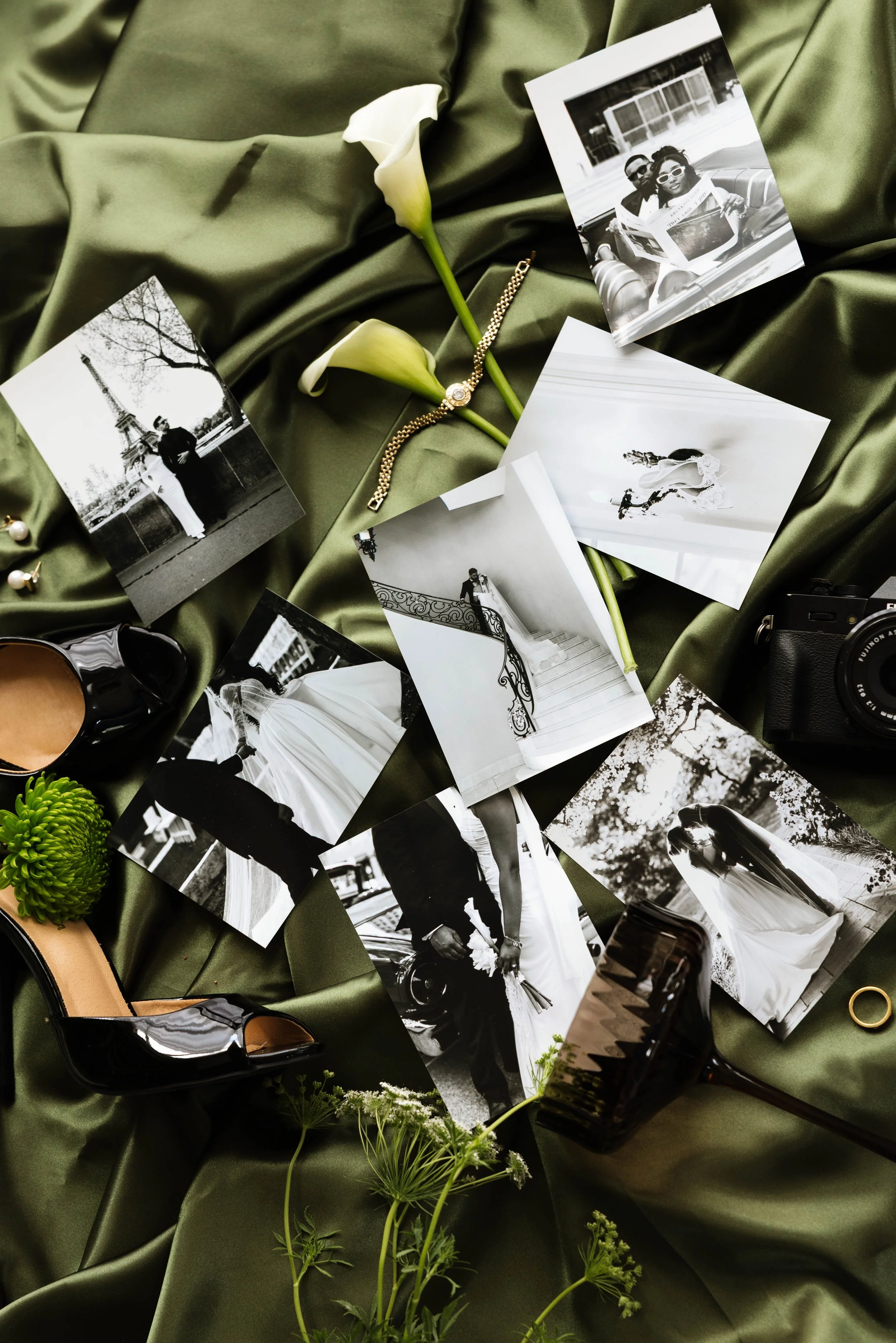 A collection of black and white wedding photographs, a gold bracelet, white calla lilies, and other small decorative items arranged on rumpled green satin fabric.