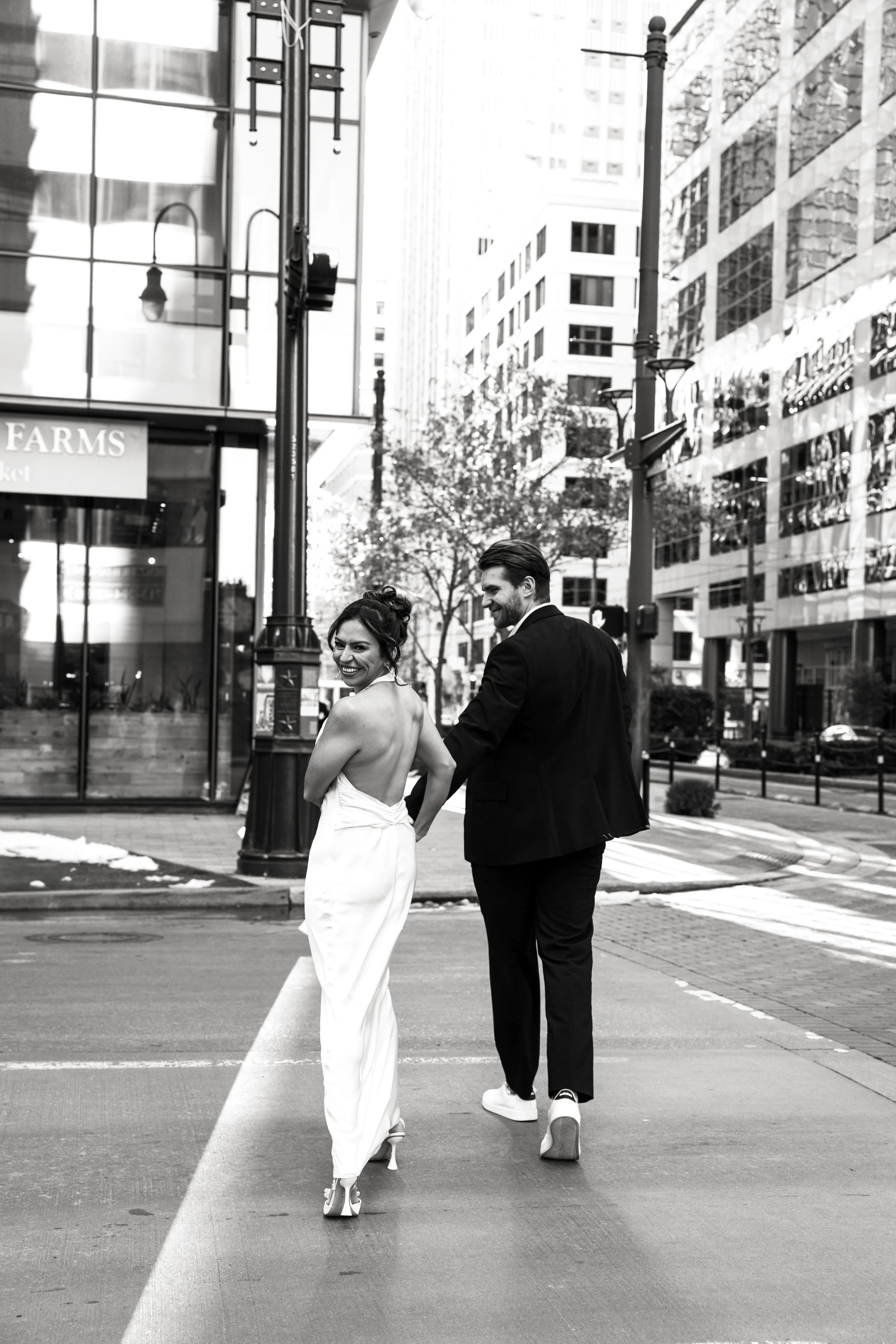 A smiling woman in a white dress and heels, and a man in a suit and sneakers, walking together on a city street.