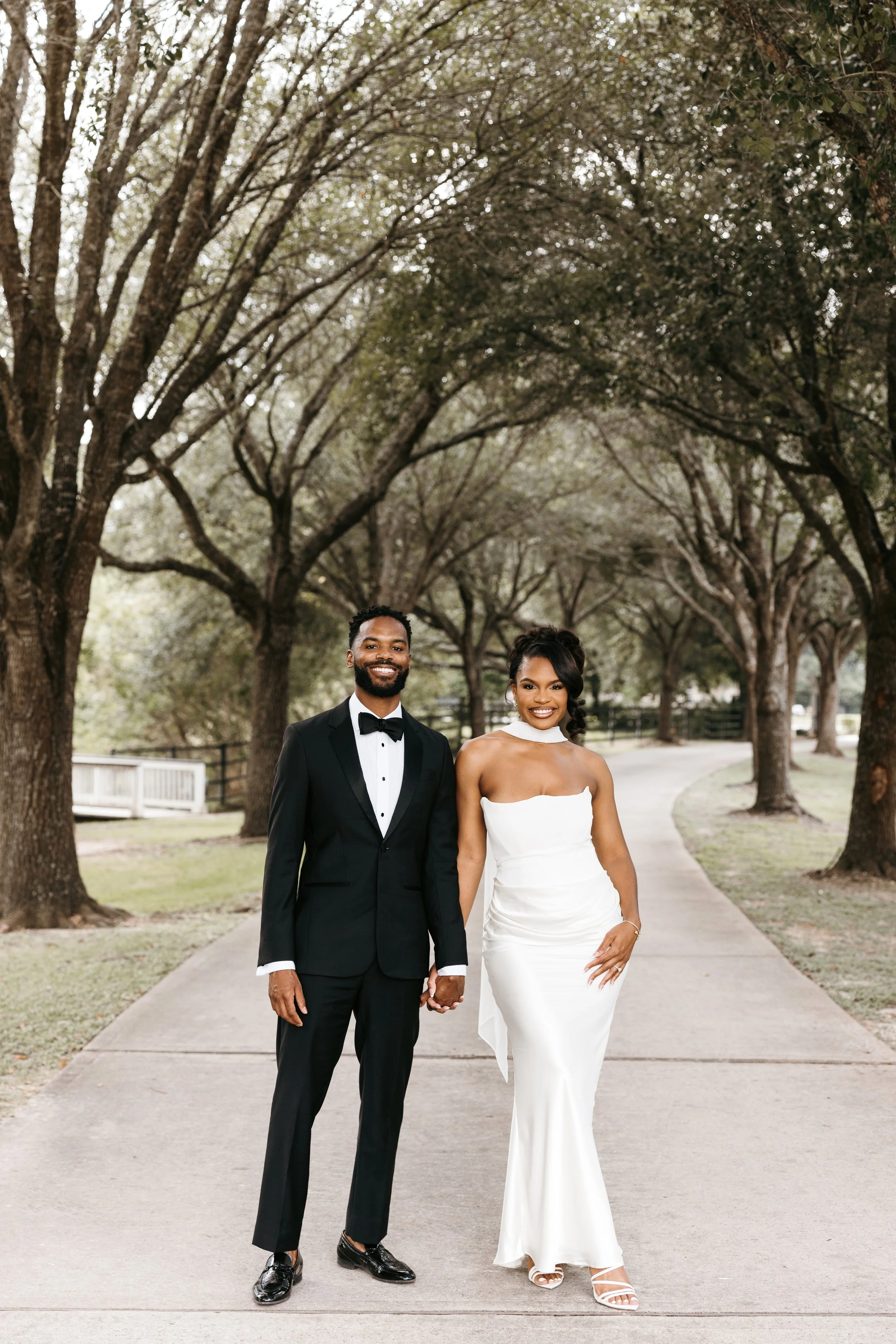 A wedding couple walking hand in hand on a tree-lined sidewalk. The groom is wearing a black tuxedo with a bow tie, and the bride is wearing a strapless white wedding gown with matching heels.