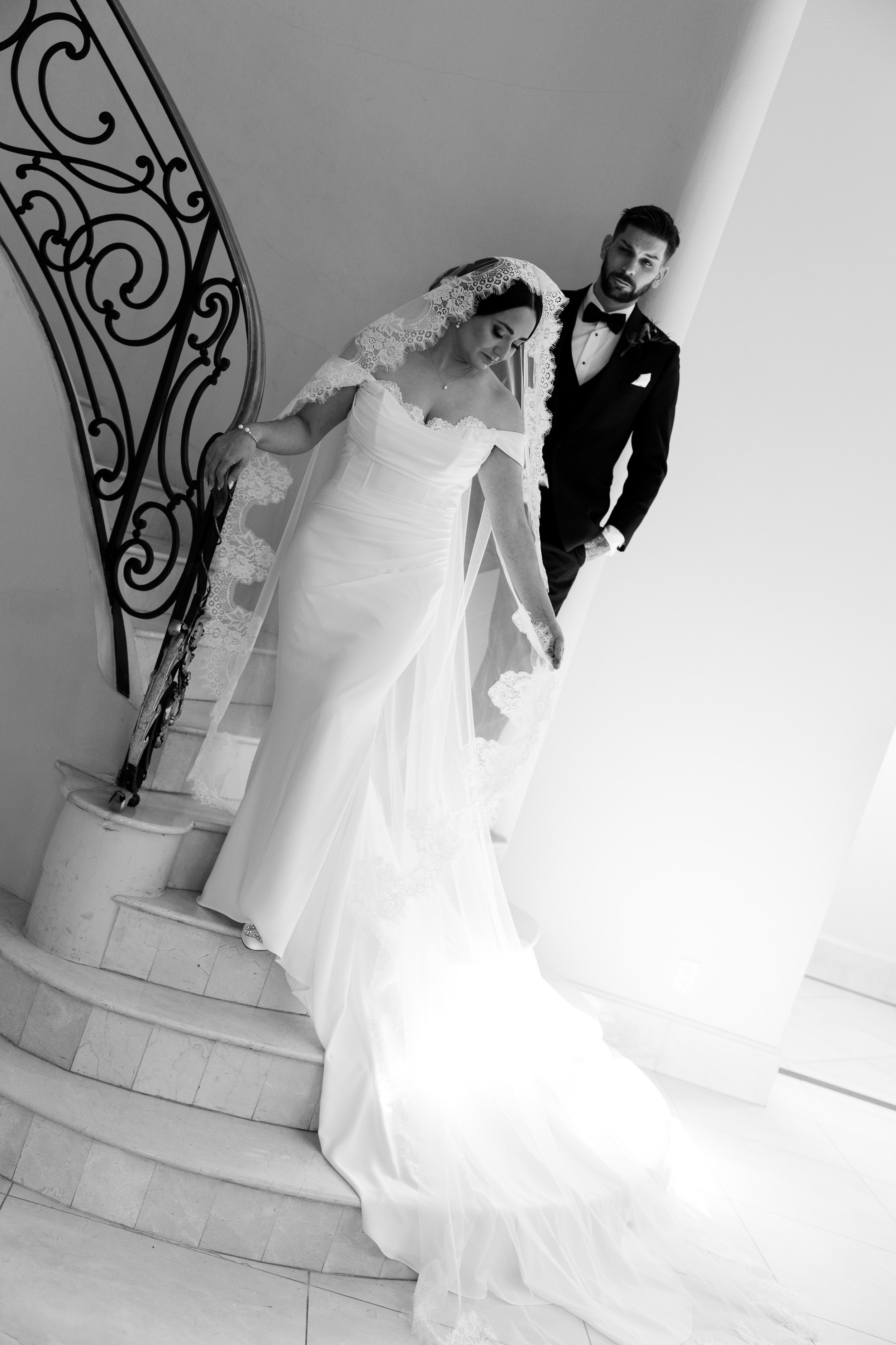 Black and white photo of a bride and groom on a staircase, the bride in a white dress with lace details and a lace veil, the groom in a tuxedo with a bow tie, both looking serious and composed.