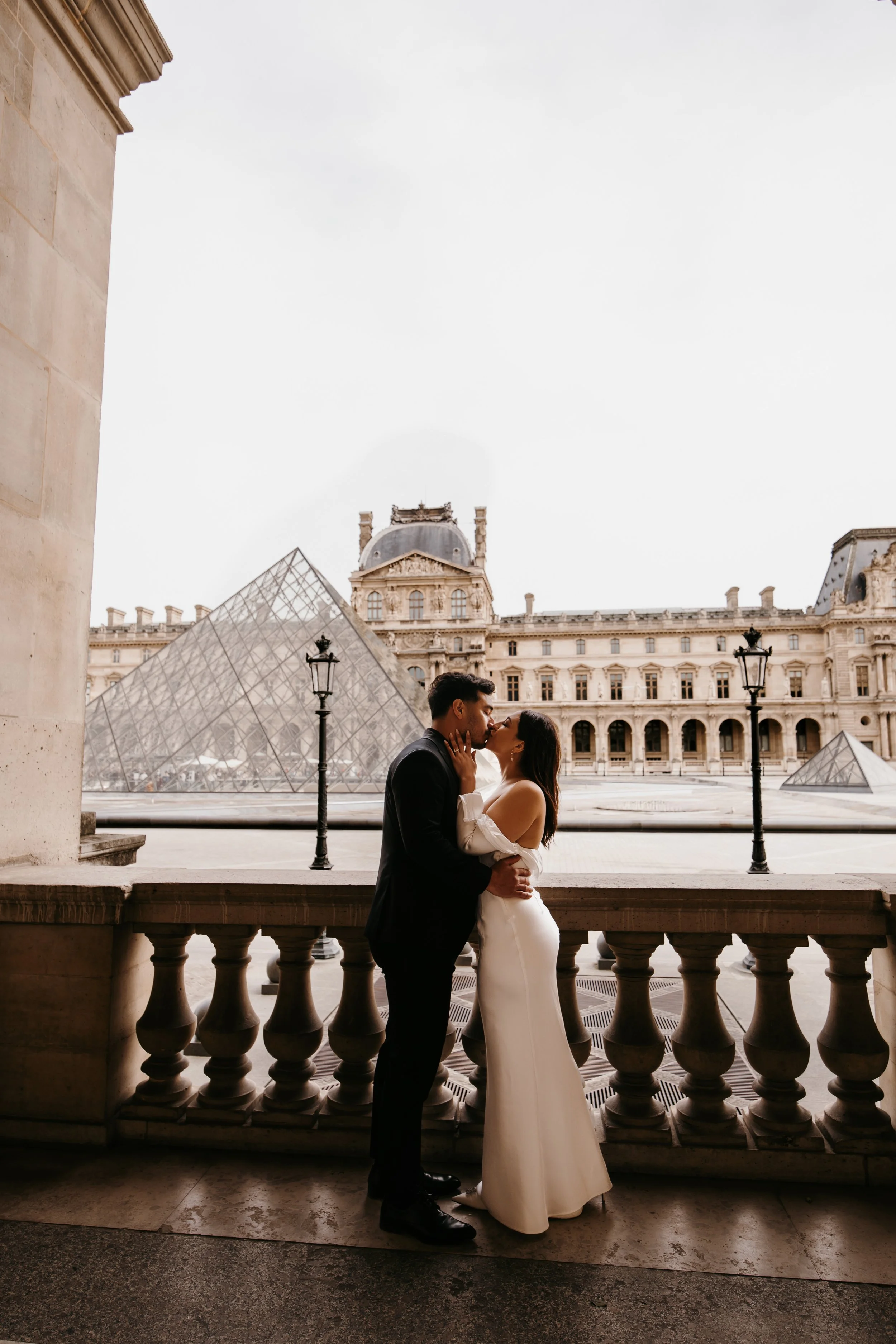 A couple in wedding attire sharing a kiss on a balcony at the Louvre Museum in Paris, with the Louvre Pyramid visible in the background.