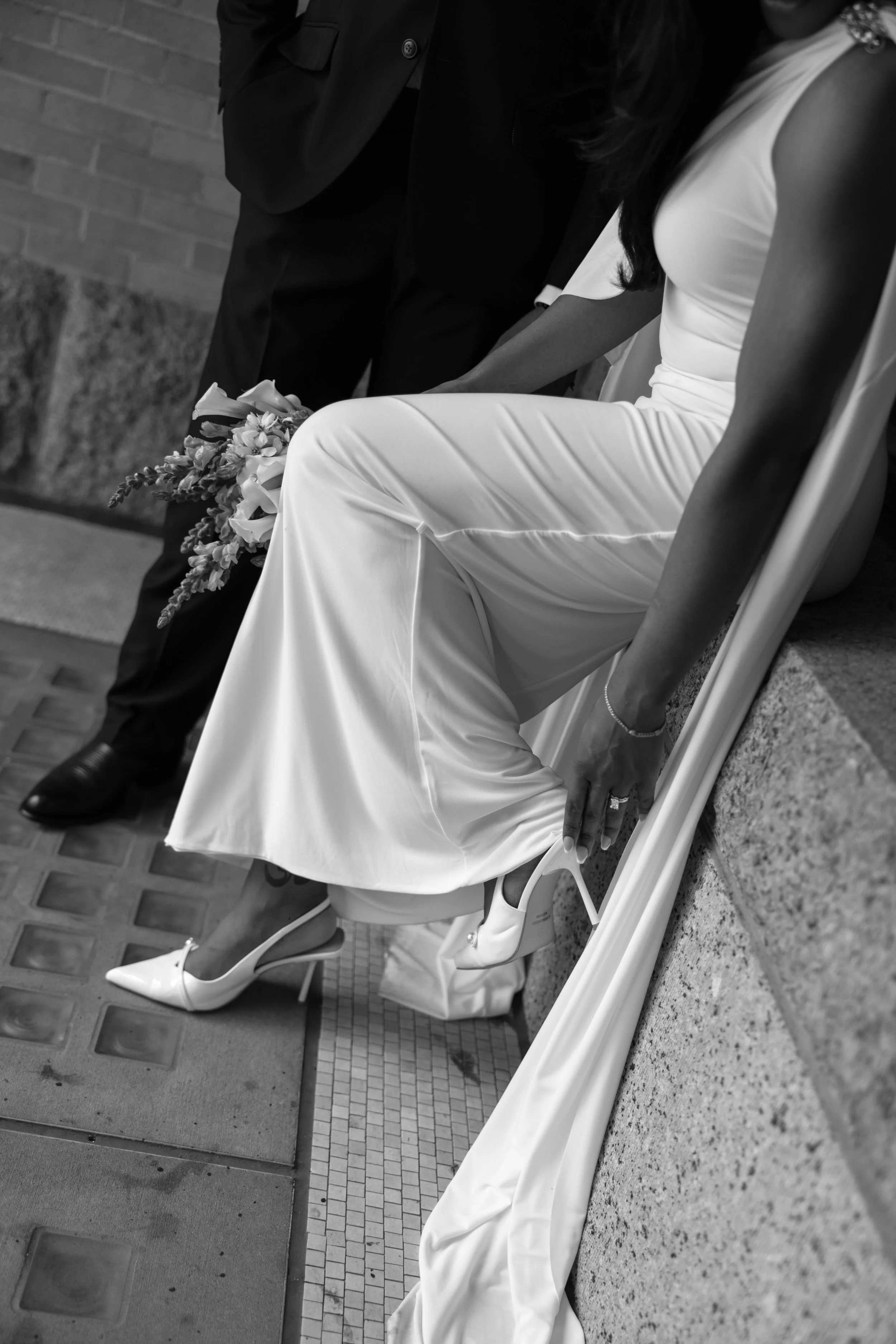Black and white photo of a woman in a white dress with a slit, sitting on a chair, with a person standing beside her holding a bouquet. The woman is wearing white heels and has a bracelet on her wrist. The person beside her is wearing a dark suit. Th
