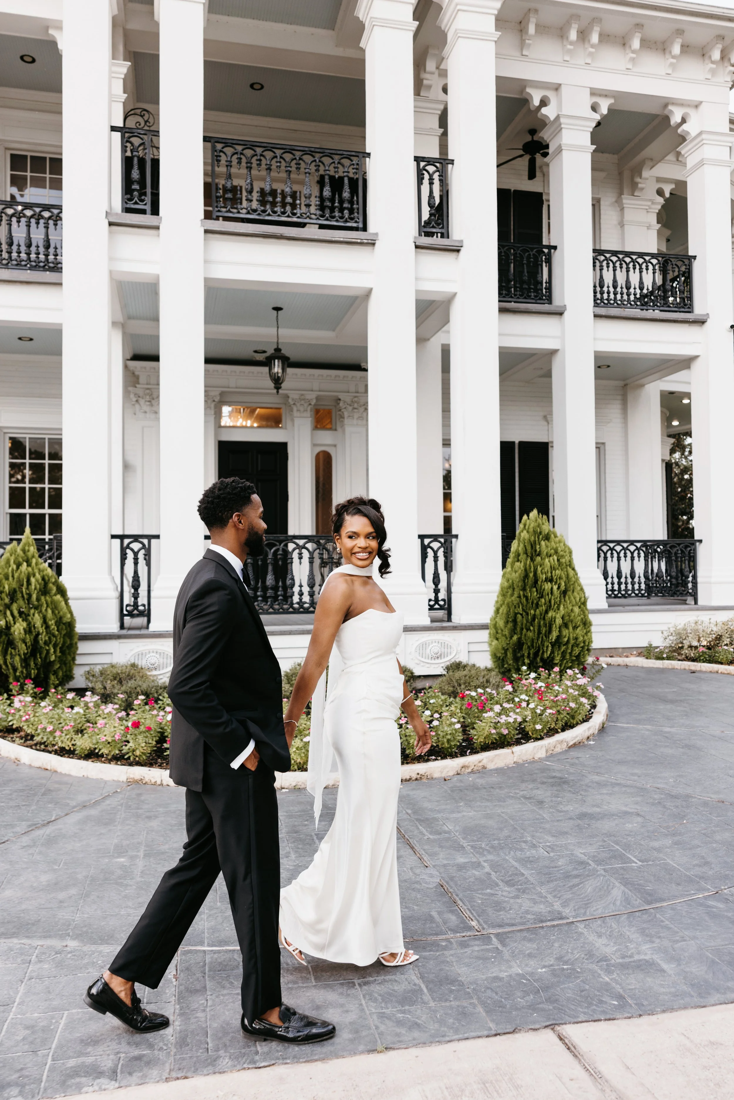 A smiling woman in a white strapless dress and a man in a black suit walking in front of a large white house with black railings and columns, with flowers and greenery in the garden.
