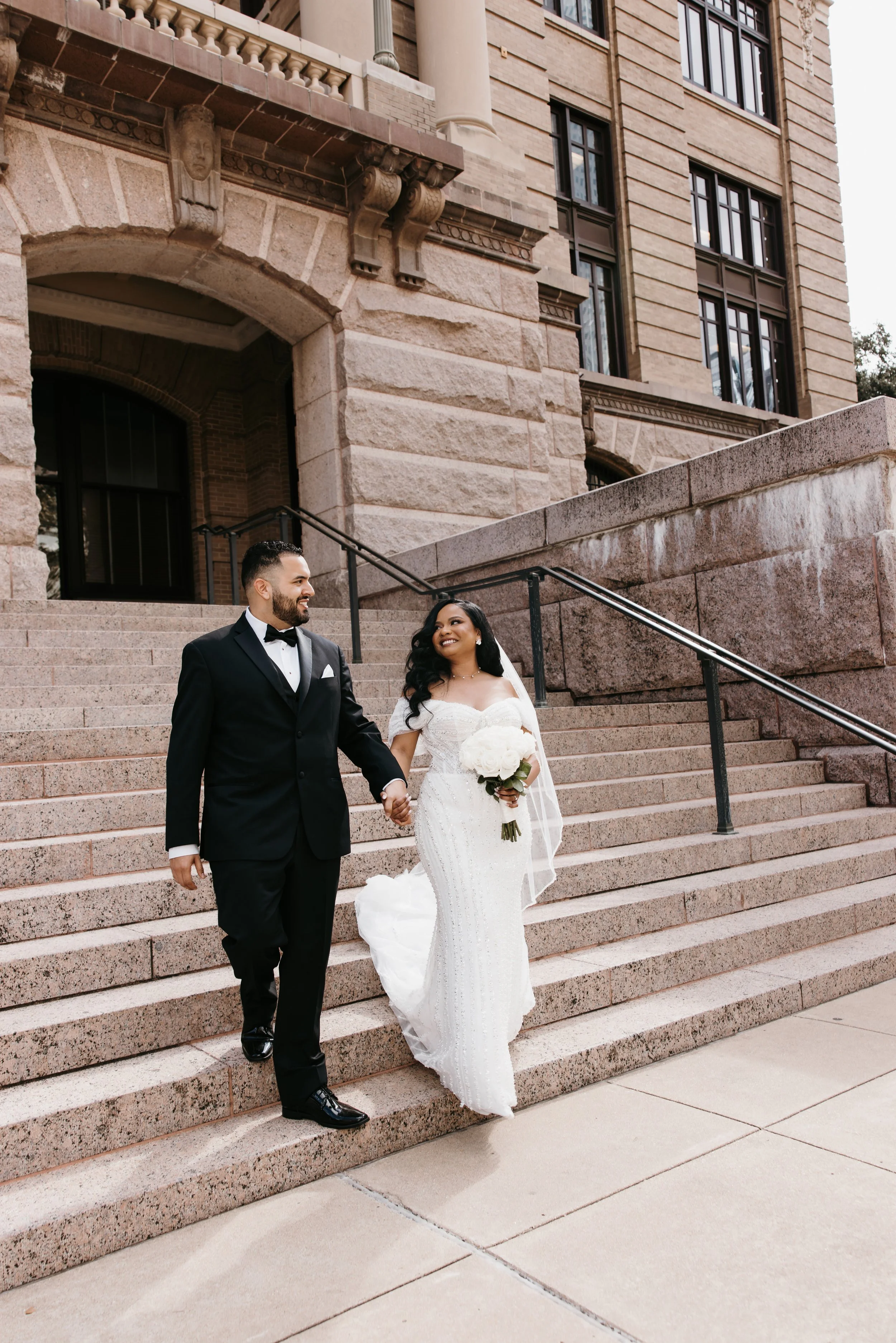 A newlywed couple walking down the steps of a historic building, holding hands, smiling, the bride in a white wedding dress holding a bouquet, and the groom in a black tuxedo with a bowtie.