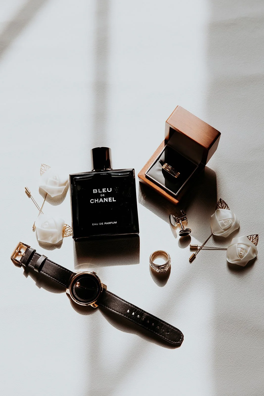 Luxury perfume, jewelry, watches, and accessories on a white surface, including a bottle of Bleu de Chanel perfume, a ring, earrings, and a black leather strap watch.