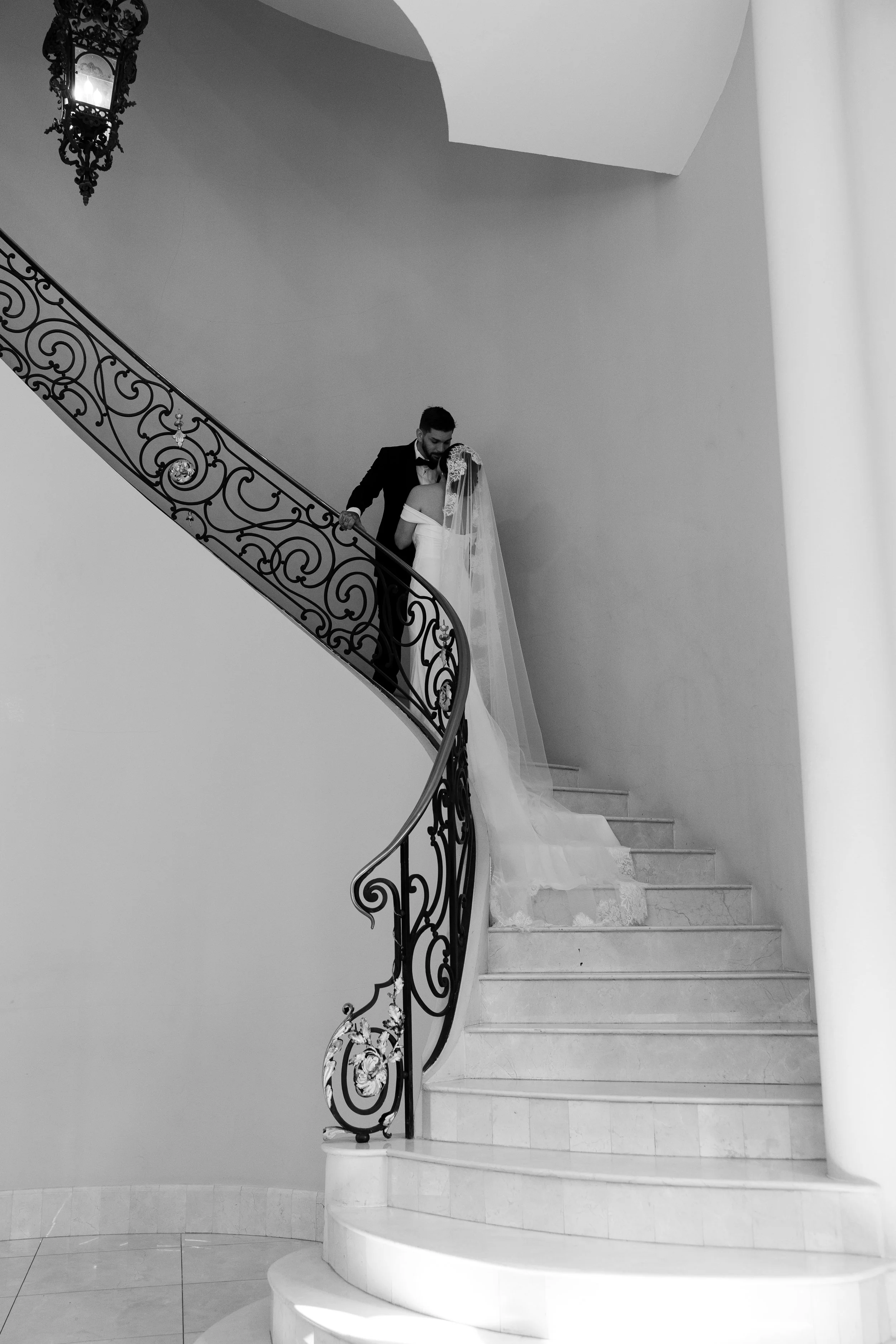 A black-and-white photo shows a man and woman standing on a grand staircase with ornate metal railing, possibly dressed for a wedding, in an elegant interior with a large column and a decorative lantern hanging from the ceiling.