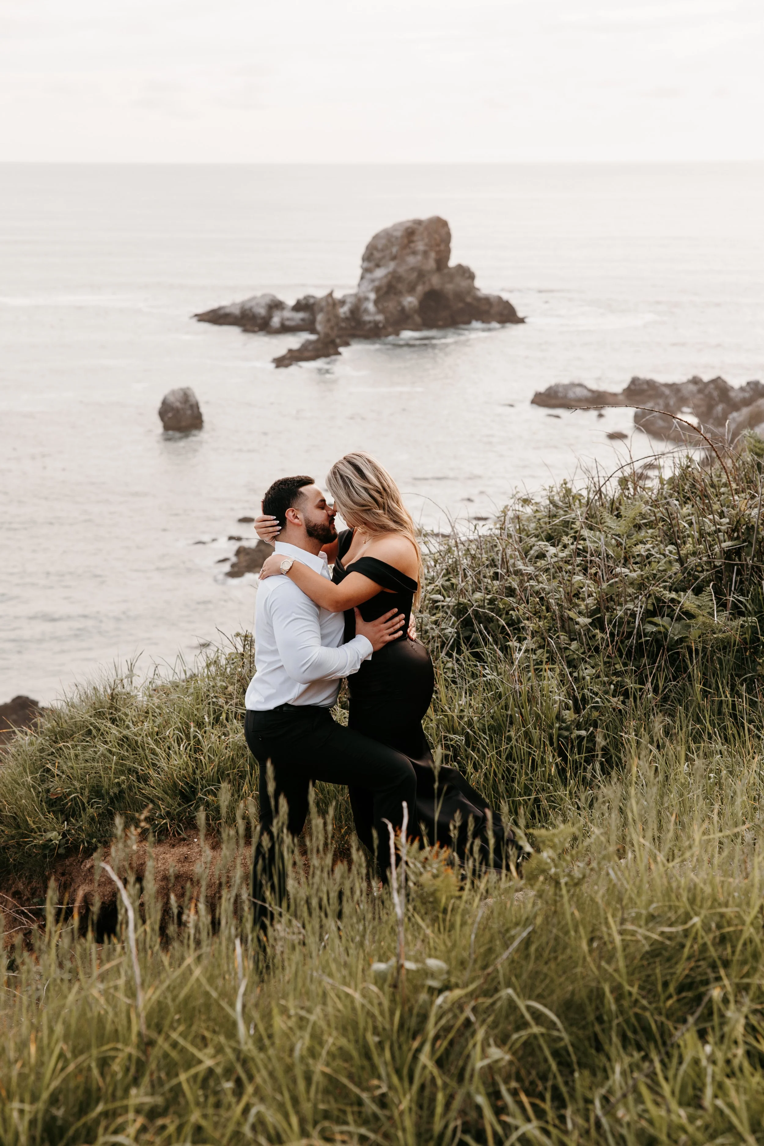 A couple embracing in a romantic kiss outdoors near the coast, with the ocean and rocks in the background.