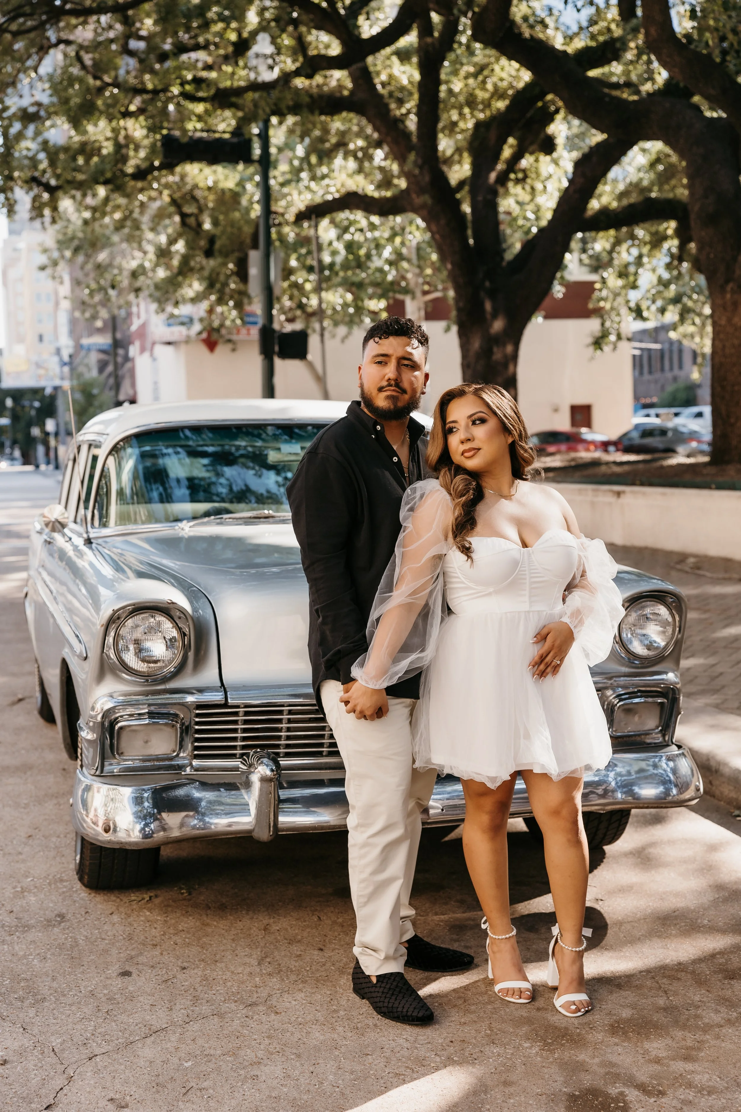 A couple stands in front of a vintage car, with the man in black shirt and beige pants, and the woman in a white dress with puffy sleeves, both holding hands and looking away from the camera. The background features trees and a city street.