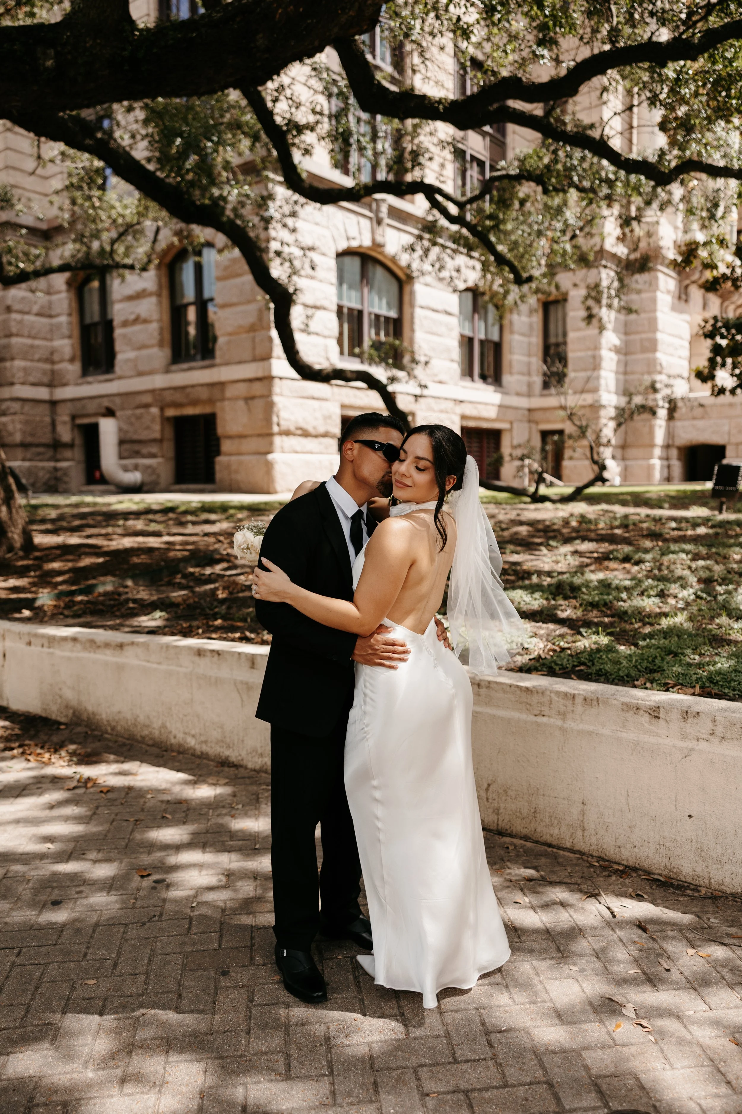 A bride and groom embrace outdoors in front of a historic brick building under a large tree with twisting branches. The bride wears a white wedding gown and veil, while the groom is dressed in a black suit with sunglasses.