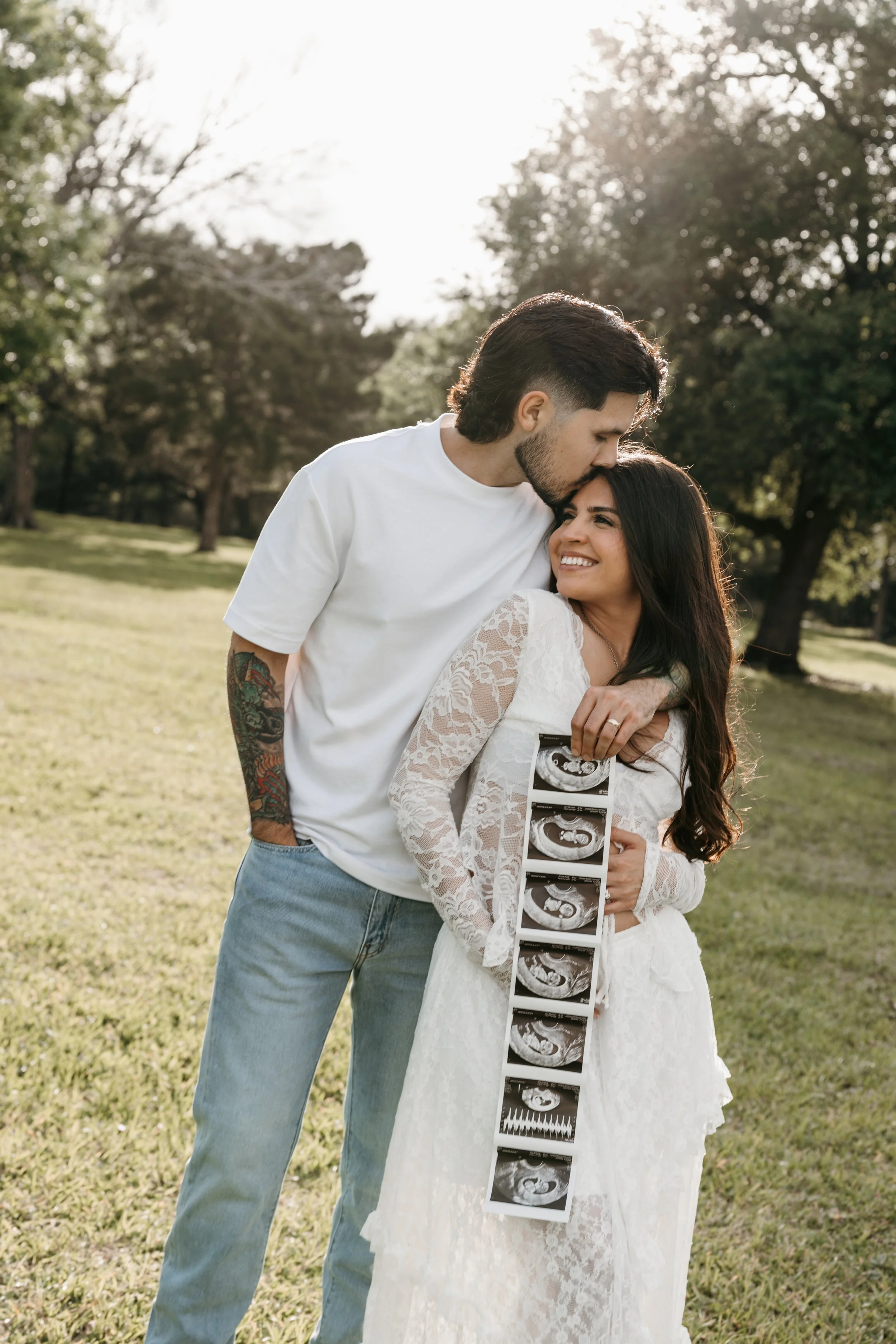 A couple stands outdoors in a park during daytime, smiling and hugging. The woman holds ultrasound images, indicating she is pregnant. The man kisses her forehead, and they are surrounded by trees with sunlight filtering through.