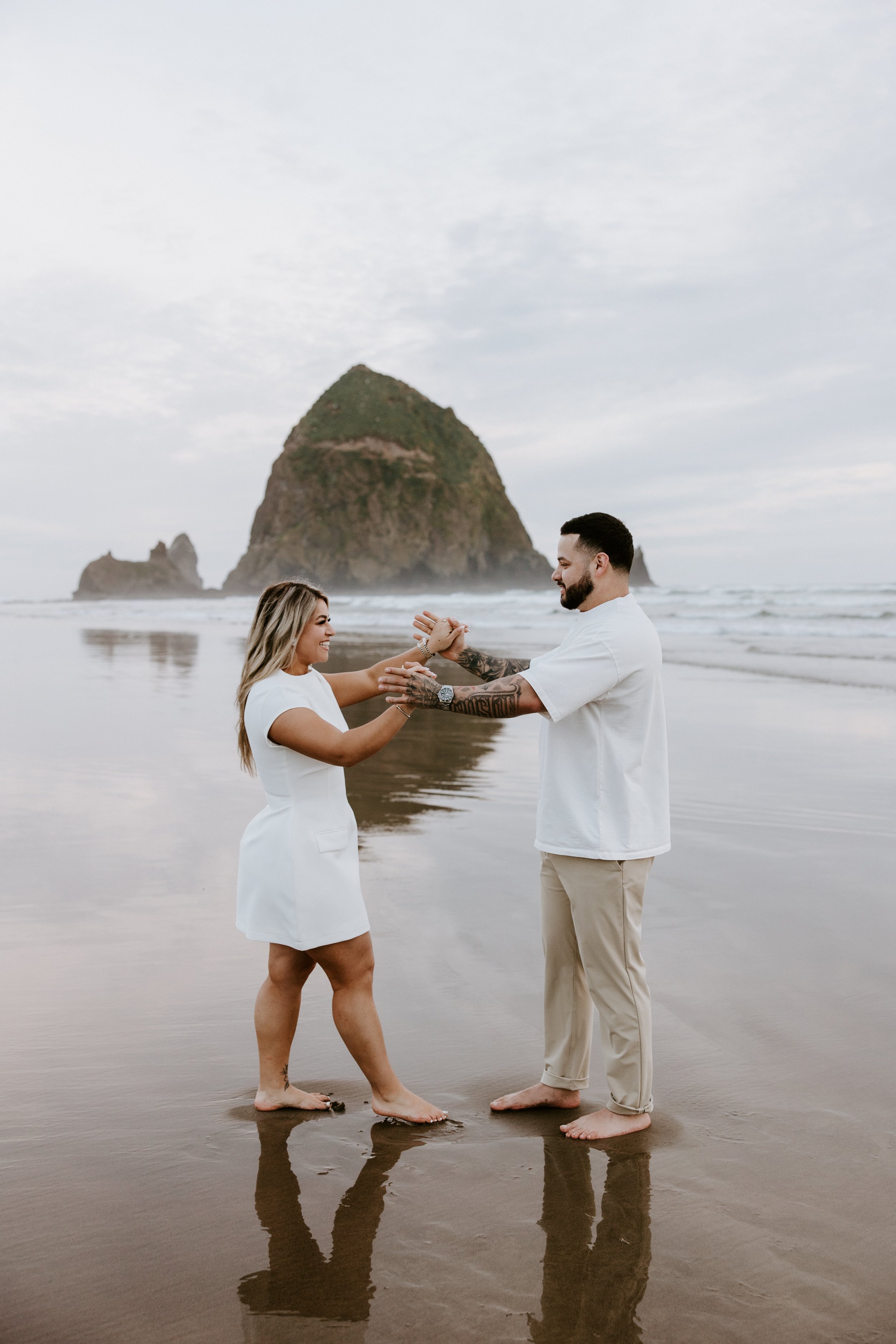 A couple stands on the beach, holding hands and smiling at each other, with a large rock formation and smaller rocks in the background, reflecting on the wet sand.