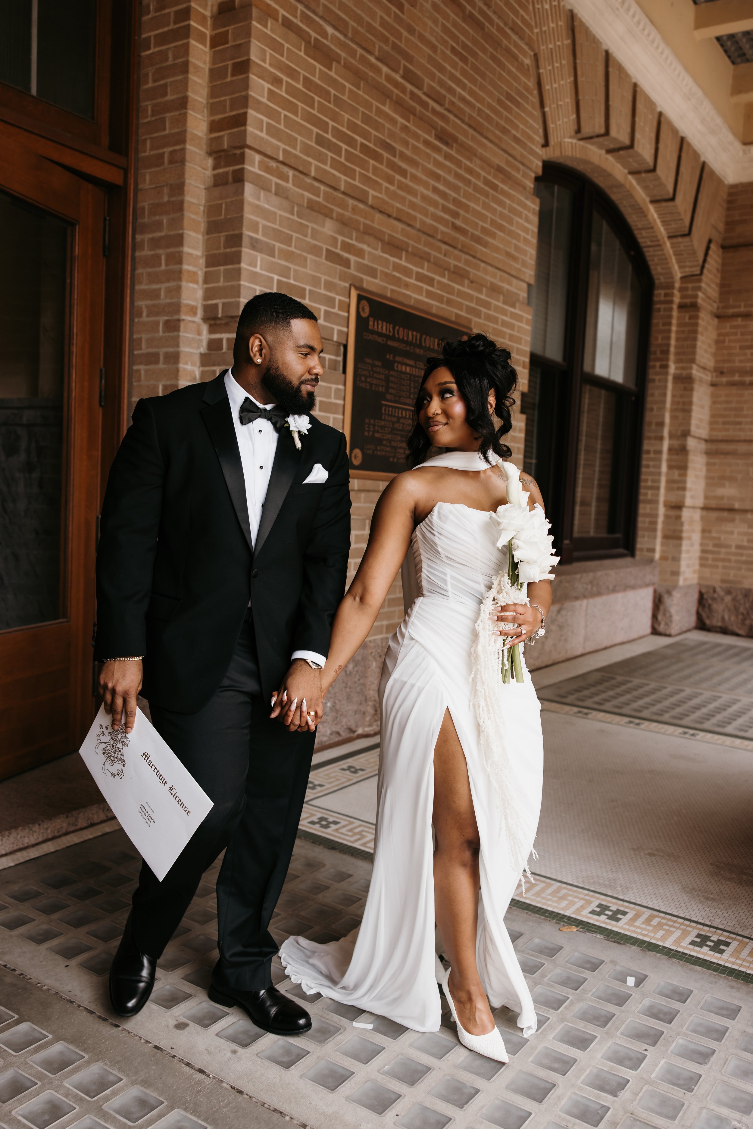 A bride and groom are standing outside a building with brick walls and large windows. The groom is wearing a black tuxedo with a bow tie and is holding a piece of paper. The bride is wearing a white wedding gown with a high slit and holding a bouquet