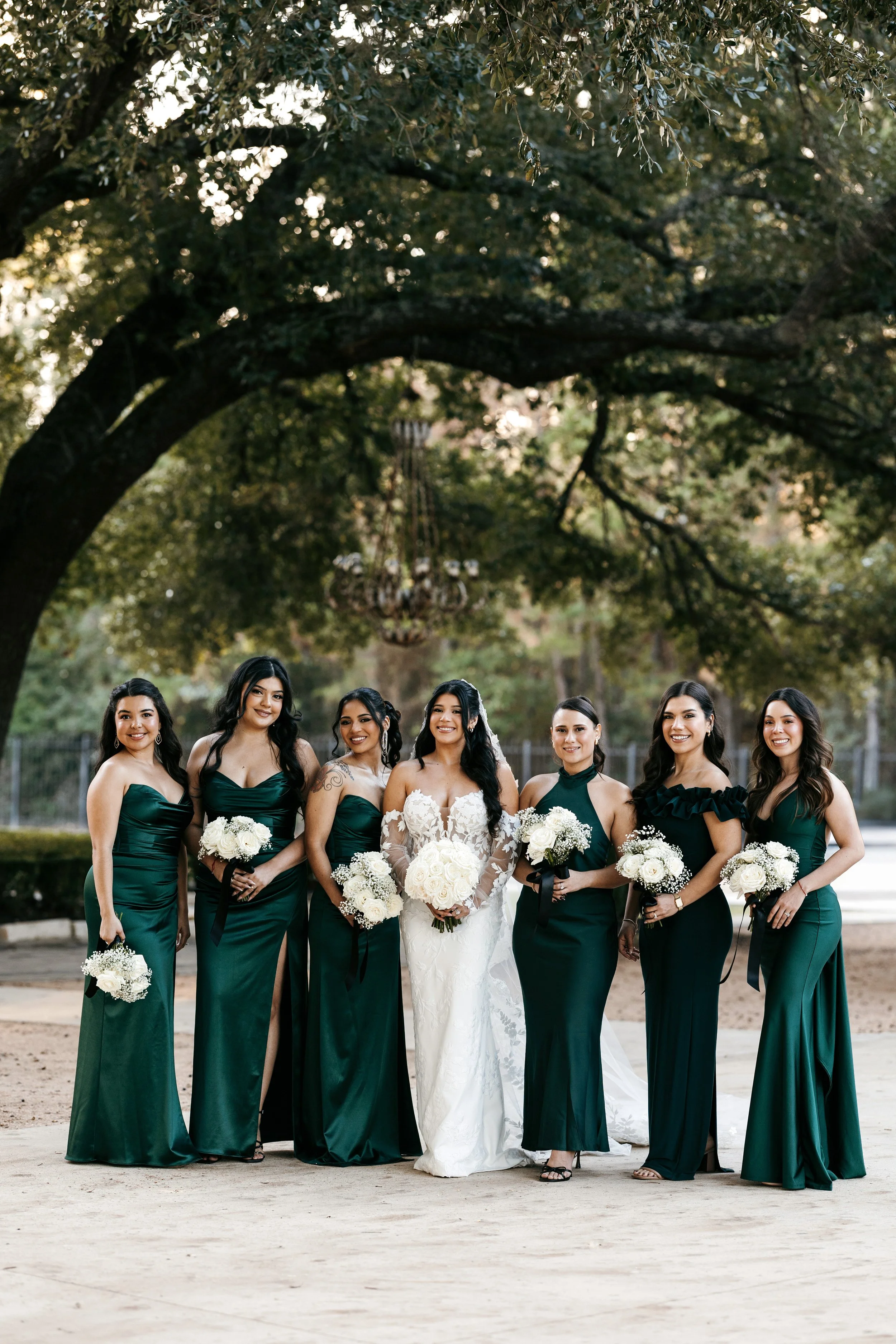 A group of eight women, including a bride in a white wedding dress, standing outdoors in front of large trees with green leaves, during the daytime. The women are dressed in matching long emerald green dresses and are holding bouquets of white flower