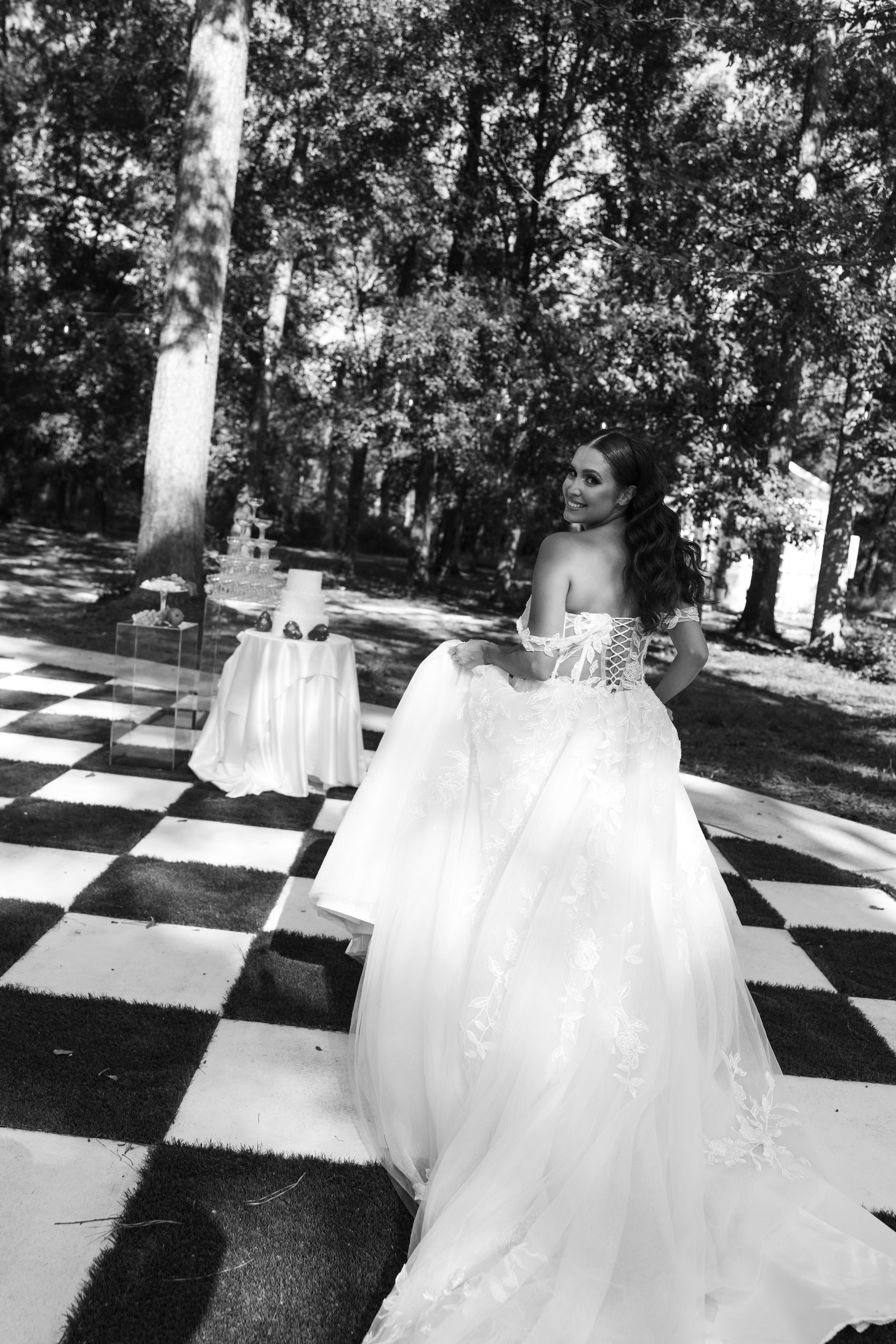 A woman in a wedding dress standing outdoors, smiling and looking over her shoulder, with tables and desserts nearby, surrounded by trees.