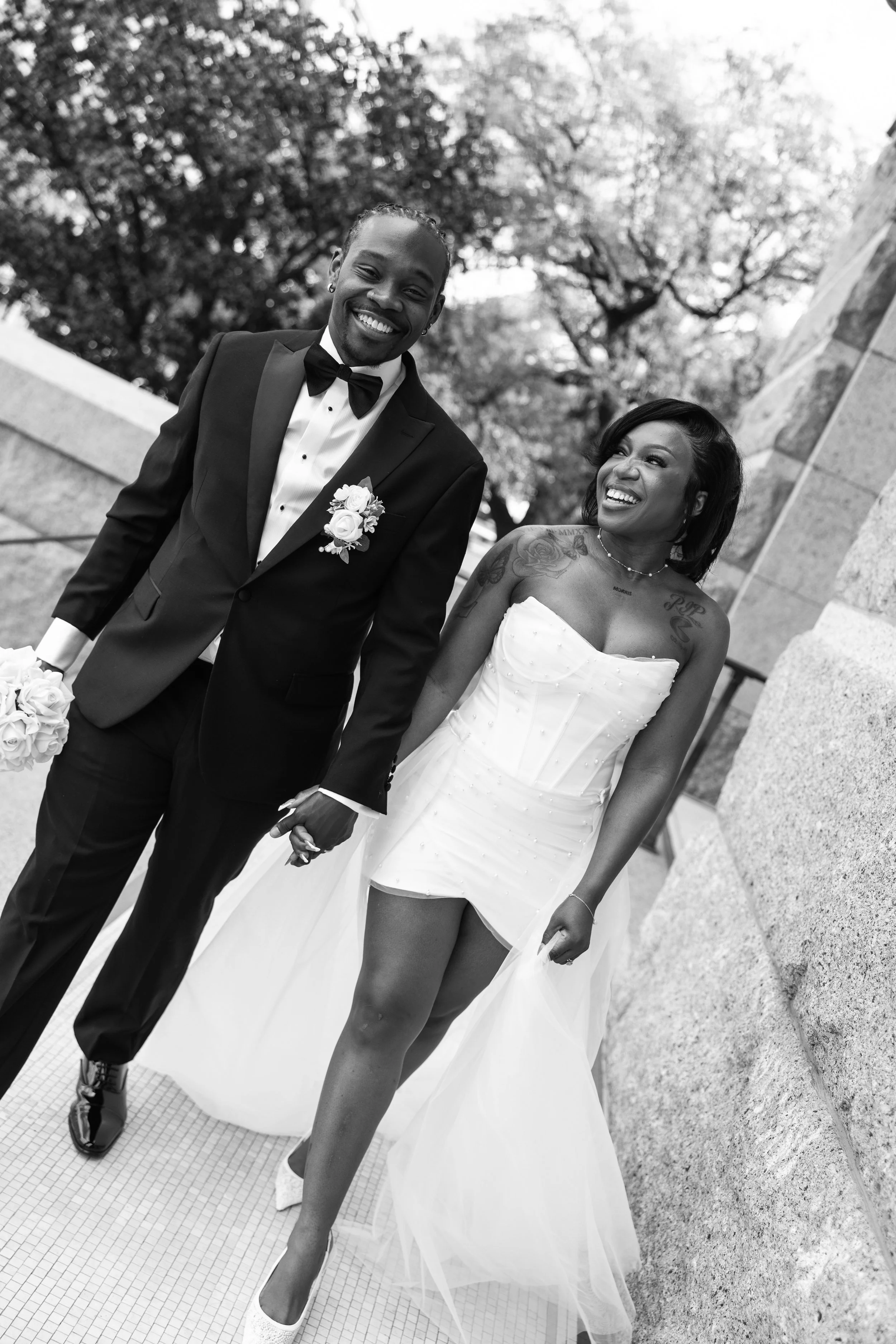 A newlywed couple holding hands and smiling outdoors. The groom is wearing a tuxedo with a bow tie, and the bride in a strapless wedding dress with tattoos visible on her shoulders and arms. Trees and a stone structure are in the background.