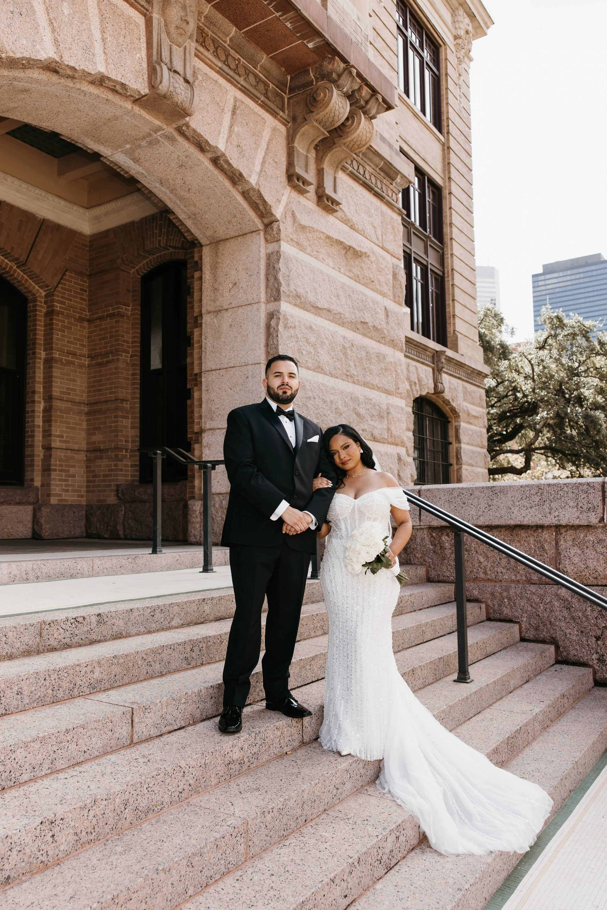 A newlywed couple standing on steps outside a historic building with ornate stonework, with skyscrapers and flowering trees in the background.