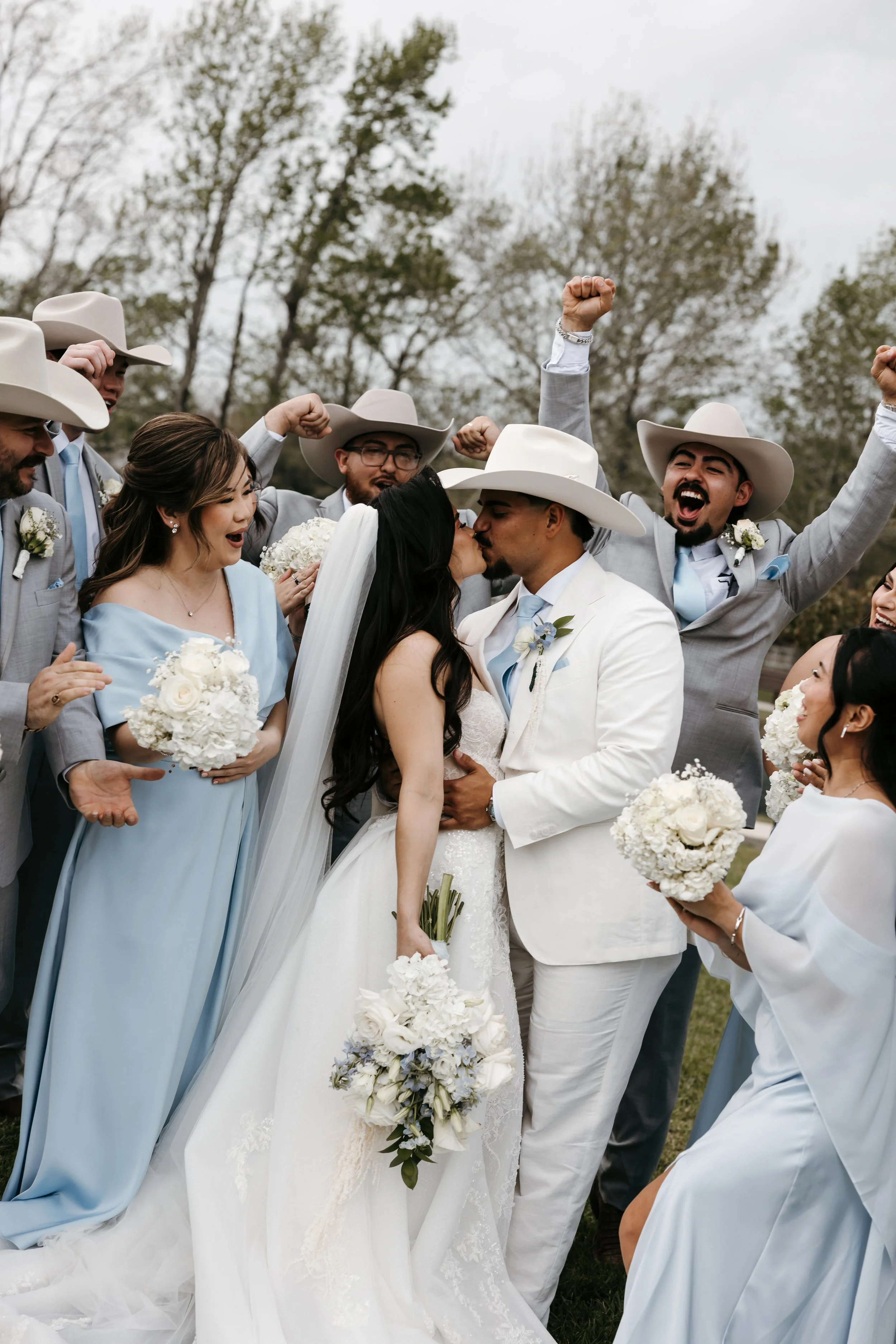 A wedding celebration with a bride and groom kissing surrounded by friends dressed in cowboy hats and suits, holding bouquets, outdoors with trees and cloudy sky in the background.