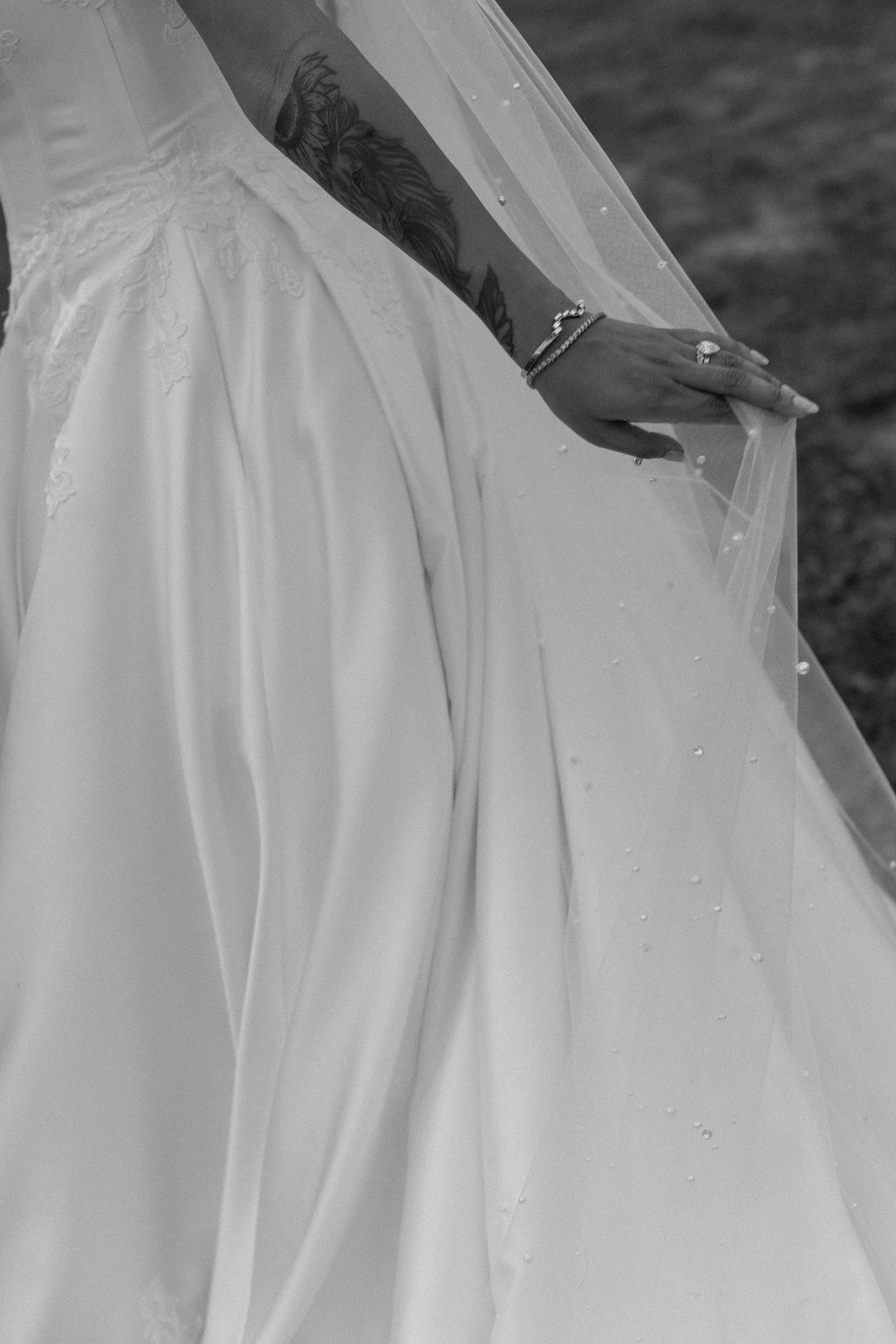 Close-up of a person wearing a wedding dress and holding a sheer veil, with a tattooed arm visible in black and white.