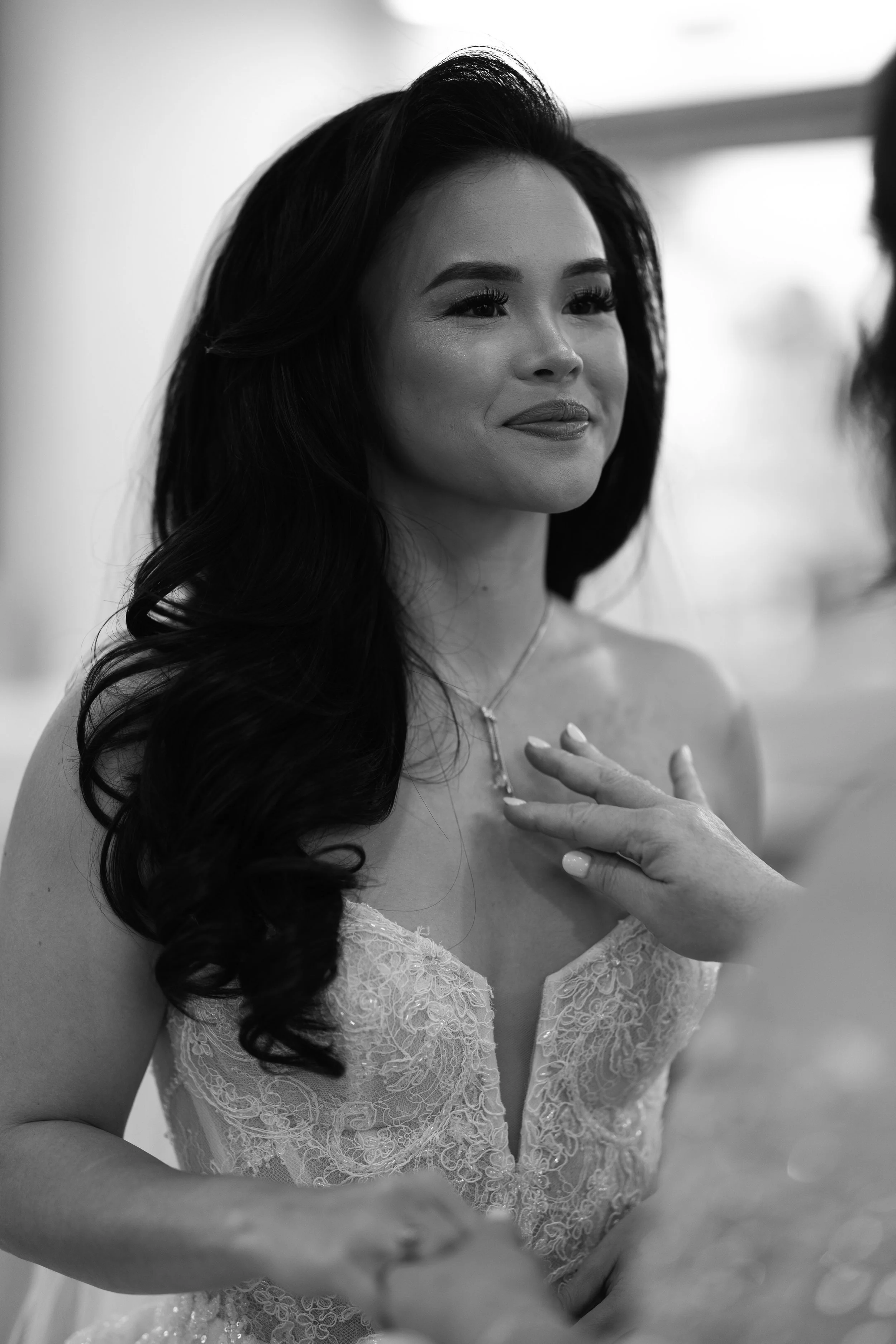 A woman with long, dark, curly hair smiling softly, wearing a strapless lace dress, with her hand on her chest, during a special moment or event.