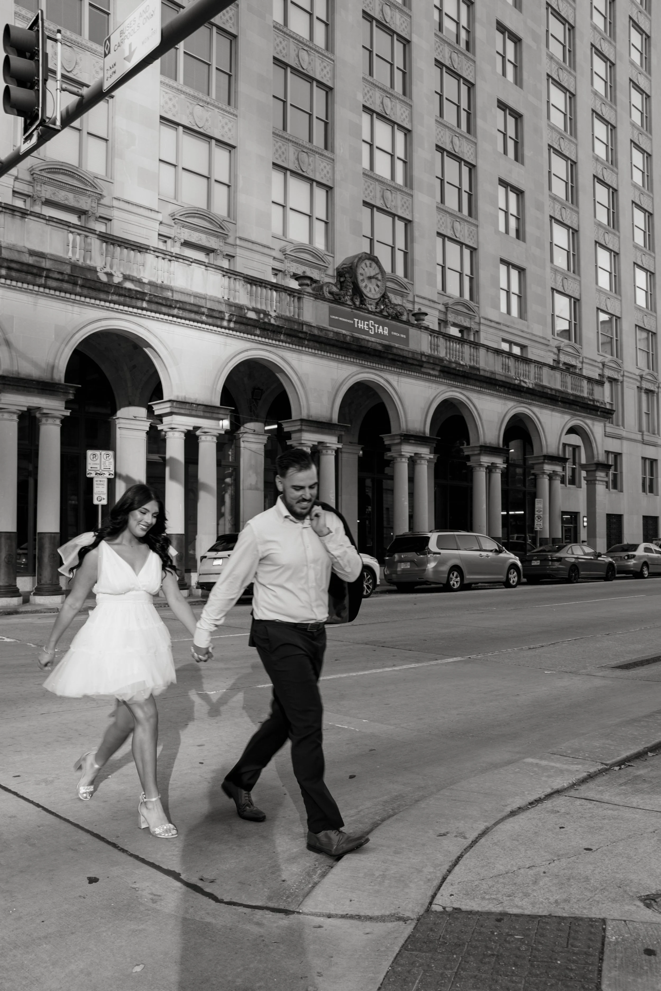 A black and white photo of a city street scene showing a woman in a white dress and heels holding hands with a man in dress clothes as they walk across the street. There are parked cars along the curb and a large building in the background with arche