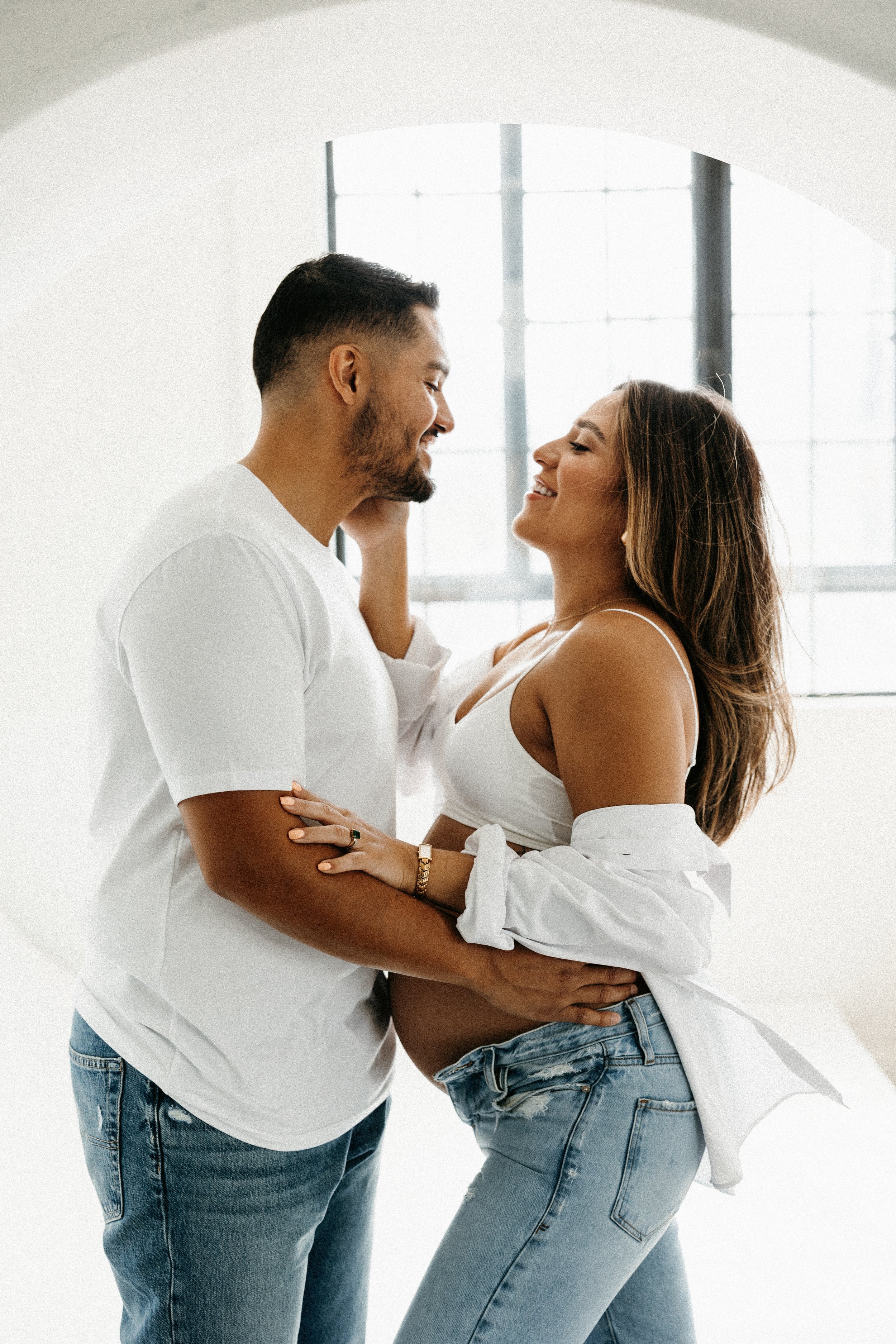 A pregnant woman and her partner sharing an intimate moment, standing close with arms wrapped around each other, inside a well-lit room with large window.