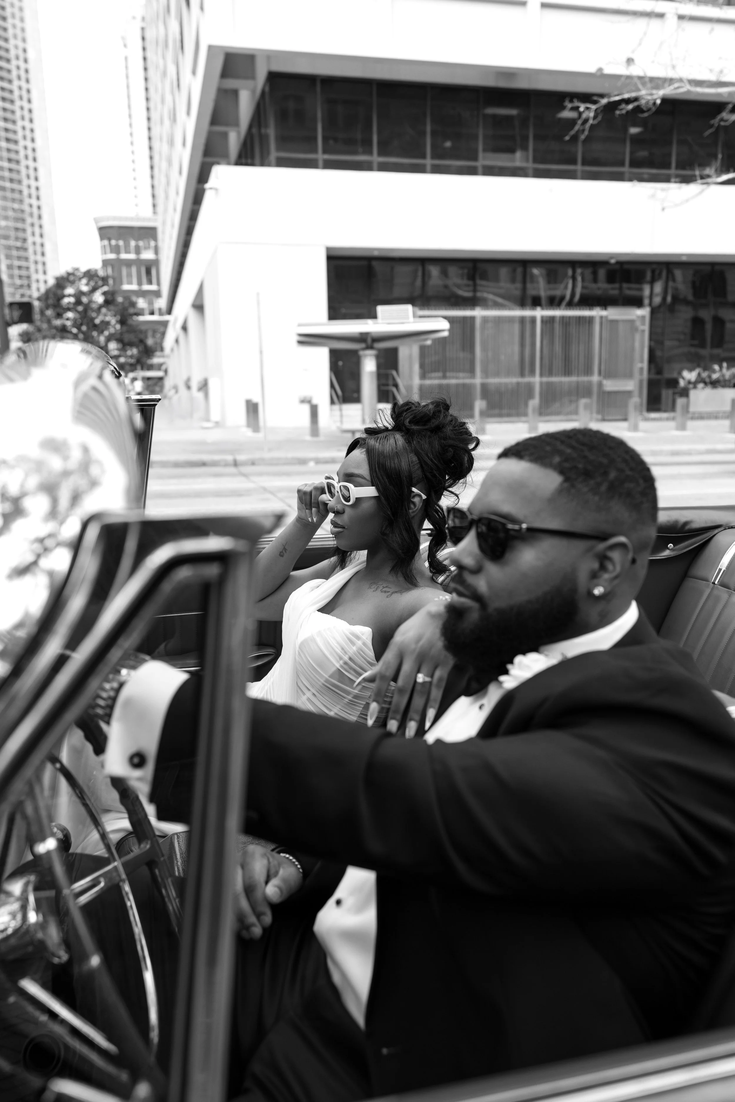 A black and white photo of a man and woman driving a convertible car in an urban area. The woman has dark hair styled in loose waves, is wearing white sunglasses, and a strapless dress. The man has short hair, a beard, and is dressed in a tuxedo with